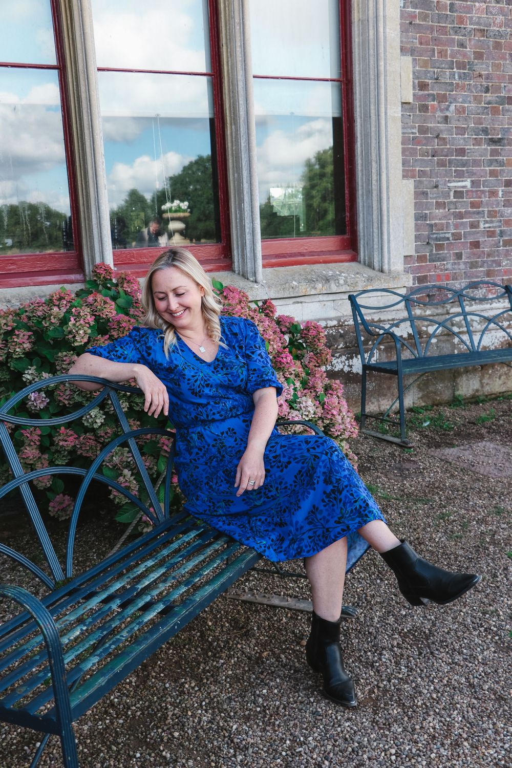 A person in a blue floral dress relaxes on a vintage teal metal bench beside pink roses and brick building.