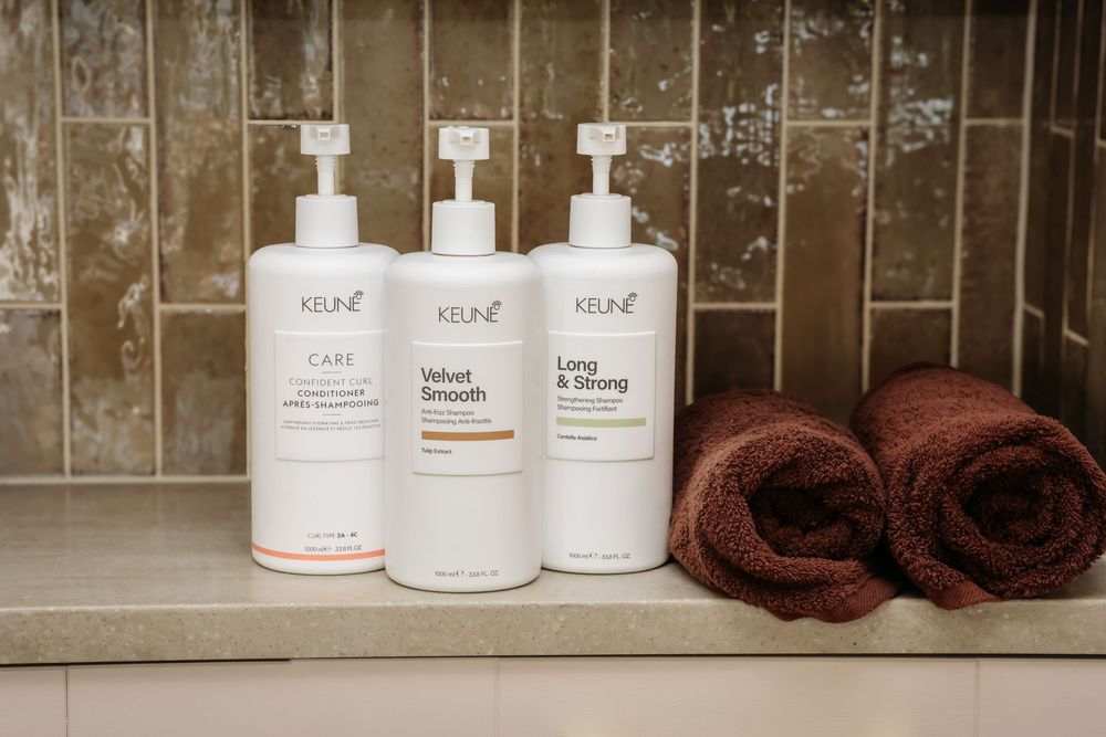Three Keune hair care bottles including Velvet Smooth and Long & Strong on a bathroom shelf with brown towels.