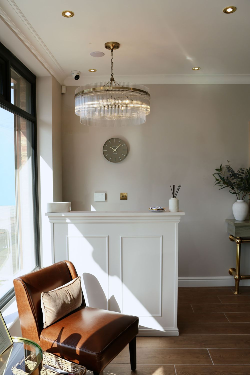 Elegant reception area with crystal chandelier, brown leather chair, white paneled desk, and large windows with ocean view.