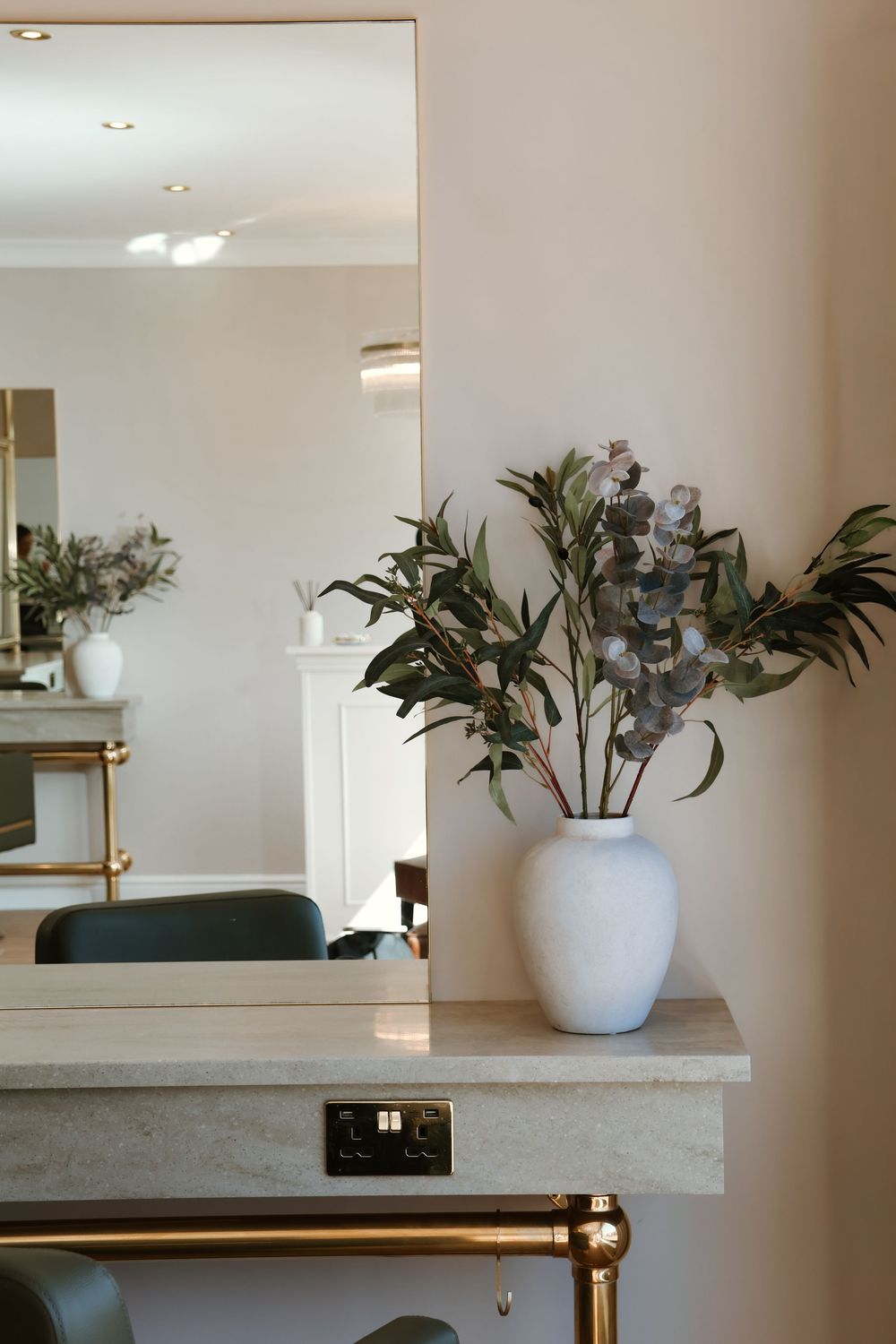 White vase with eucalyptus branches on marble vanity shelf beside large mirror in elegant salon interior.