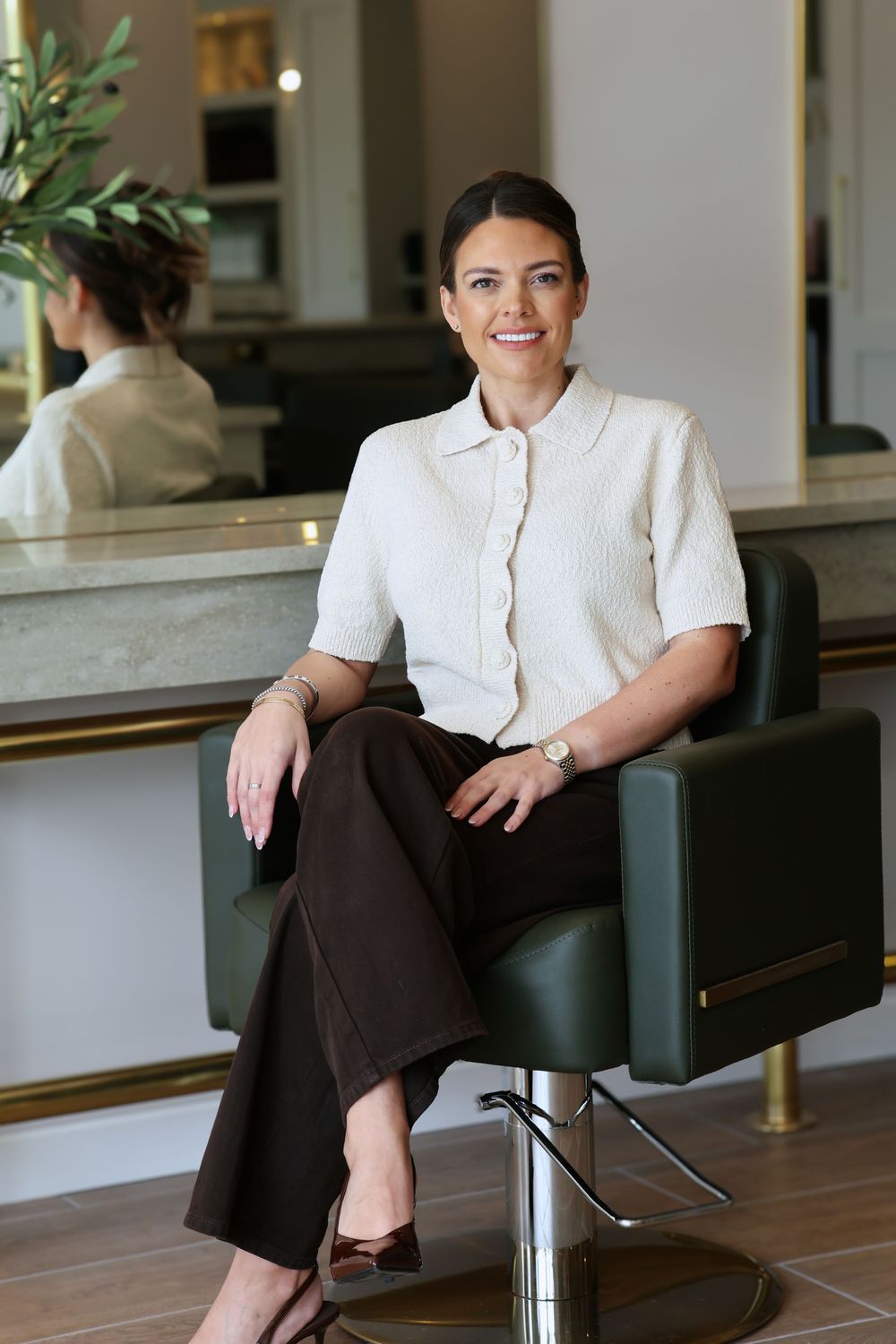 A smiling woman in a white blouse and brown trousers sits in a green salon chair in a modern hair salon.