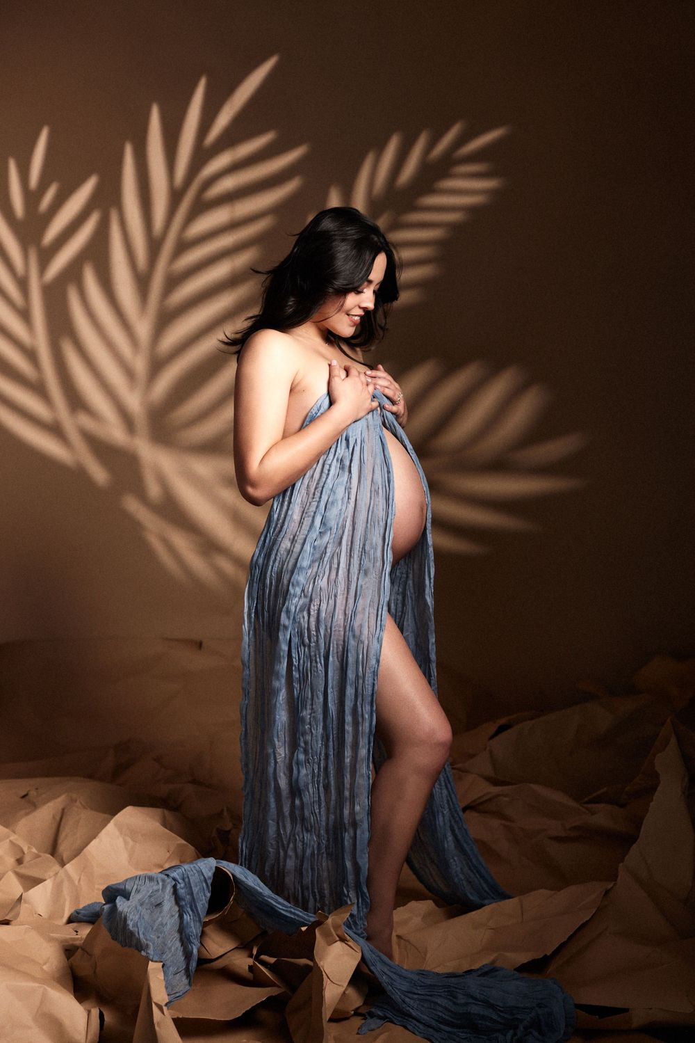 Artistic boudoir photo series with blue fabric draping and palm leaf shadow patterns against warm brown backdrop.