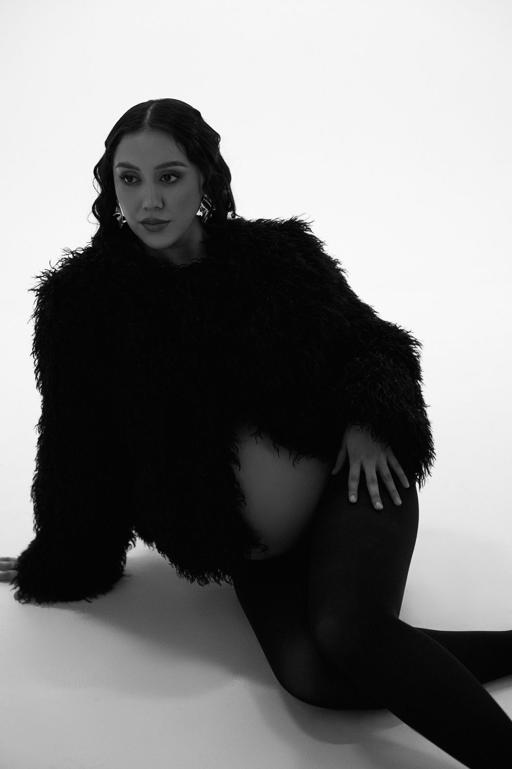 Black and white artistic portrait of a figure in a fluffy black coat posing against a white backdrop.