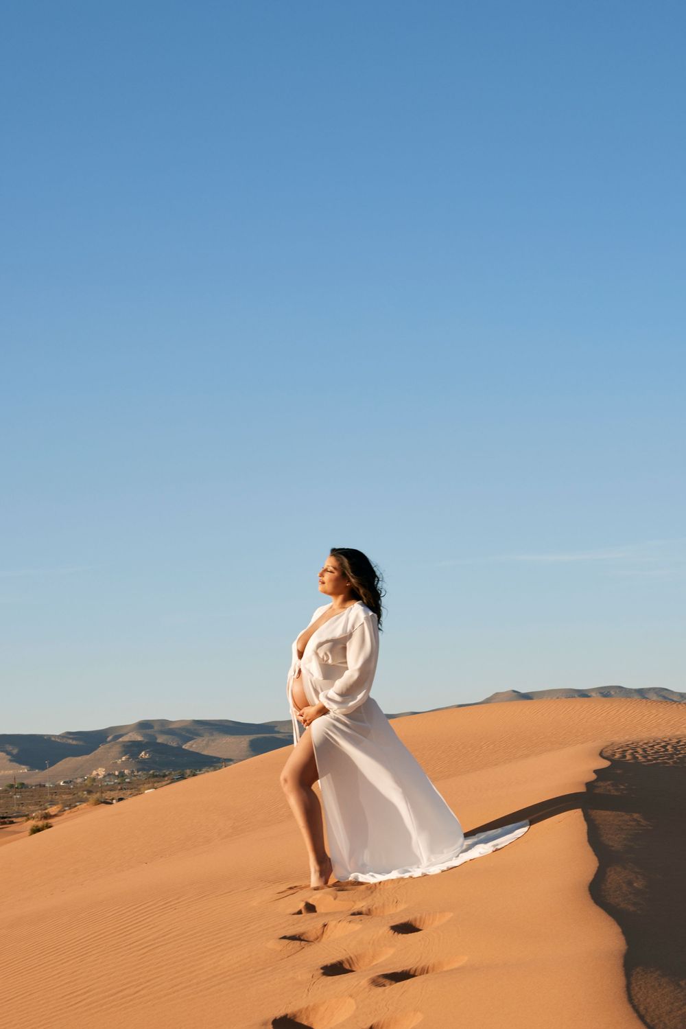 Maternity photoshoot on sand dunes with flowing white dress against blue sky.