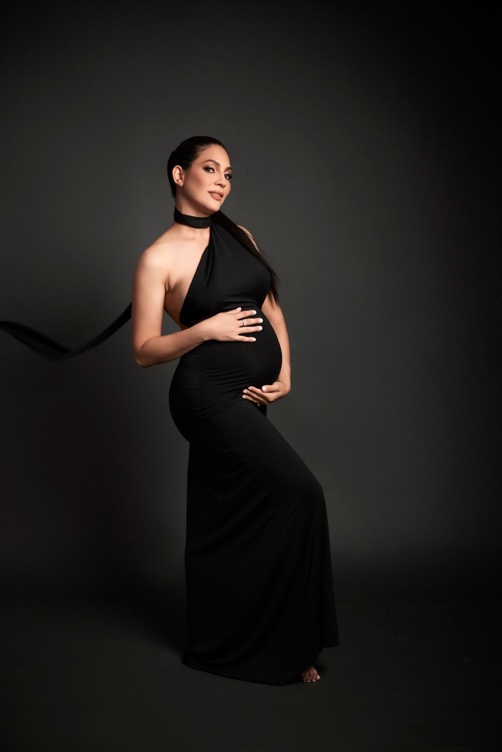 Artistic maternity portraits in black dress against dark dramatic lighting.