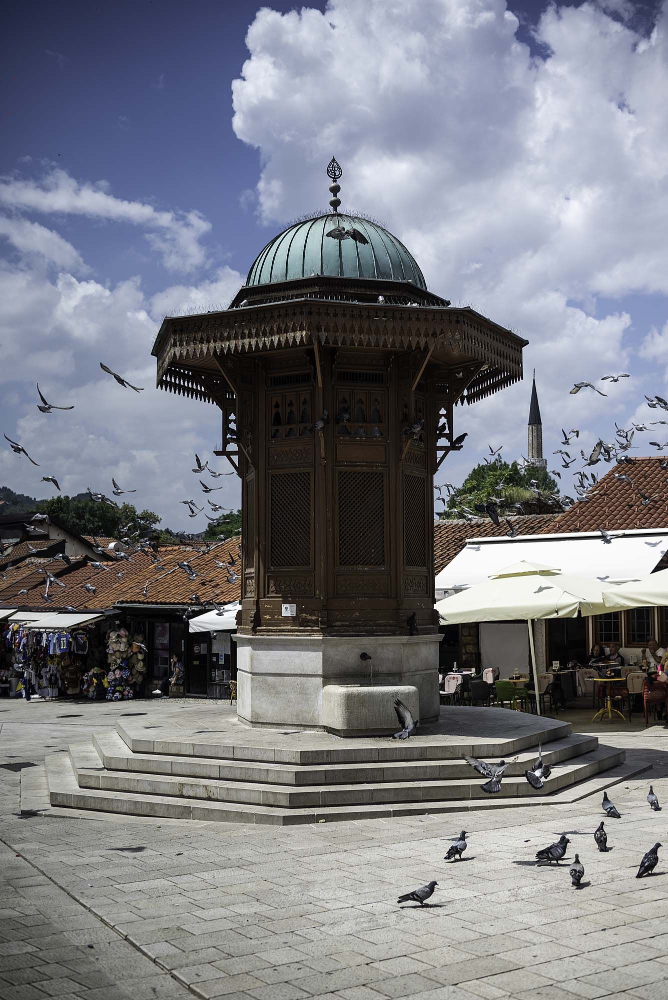 Historic gazebo in town square plaza with pigeons gathering around its stone steps under dramatic cloudy sky.