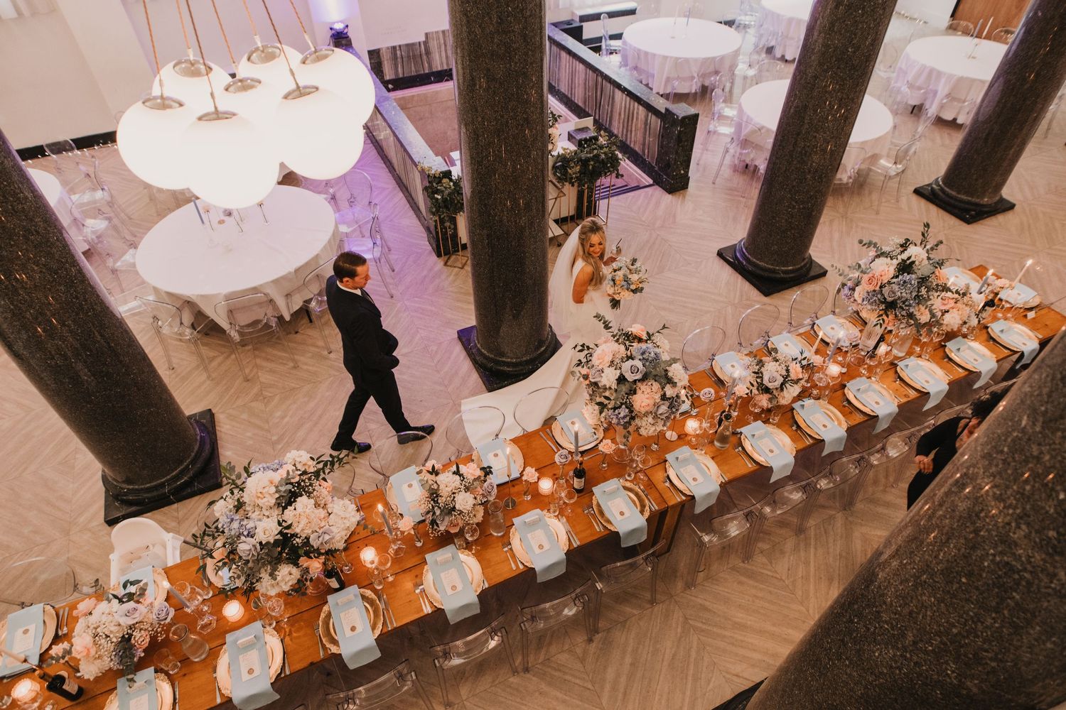 Elegant wedding reception setup with white tablecloths and floral centerpieces in a grand ballroom with columns.