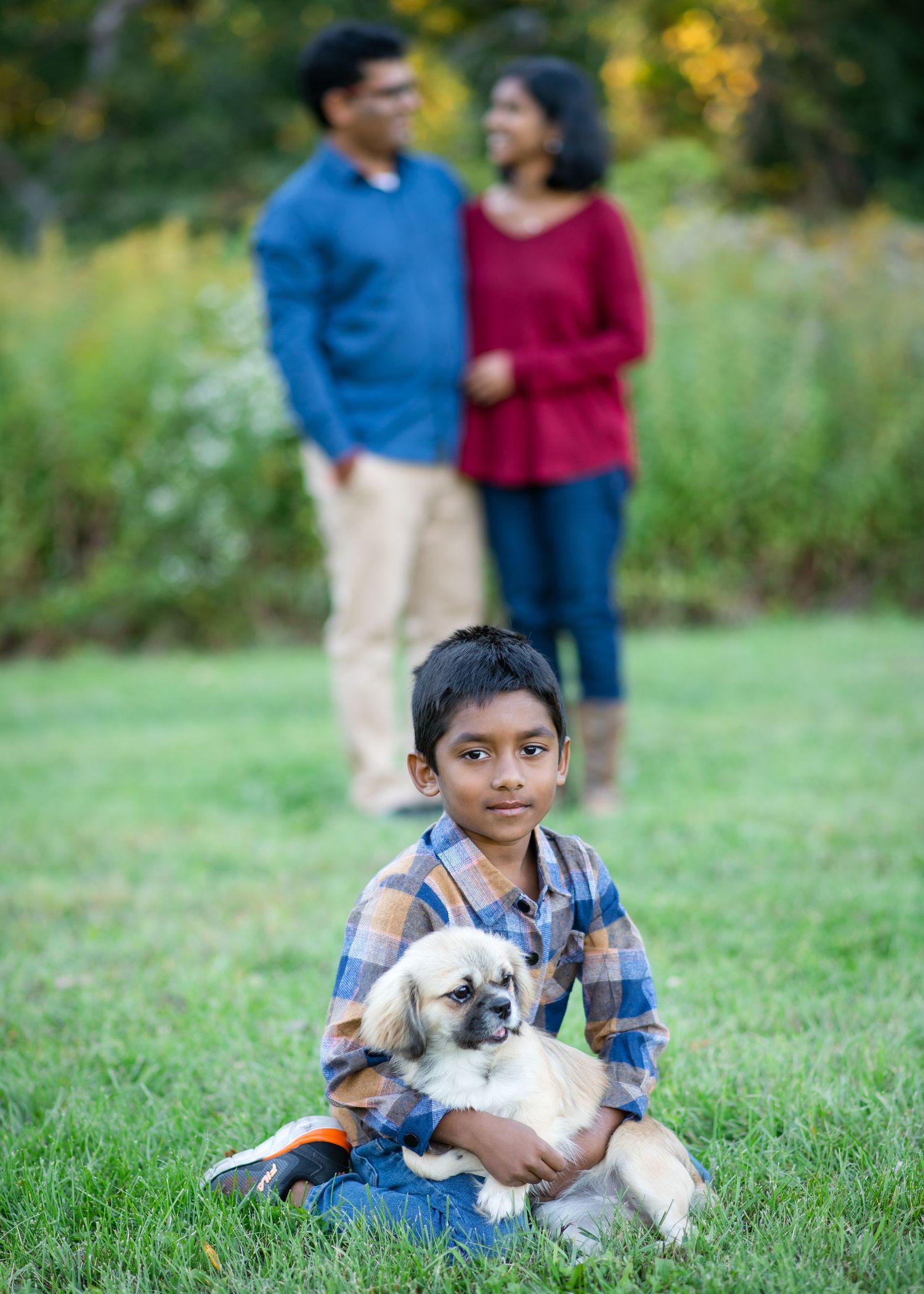 A couple holds their small brown and white dog while posing together in a sunny outdoor field at sunset.