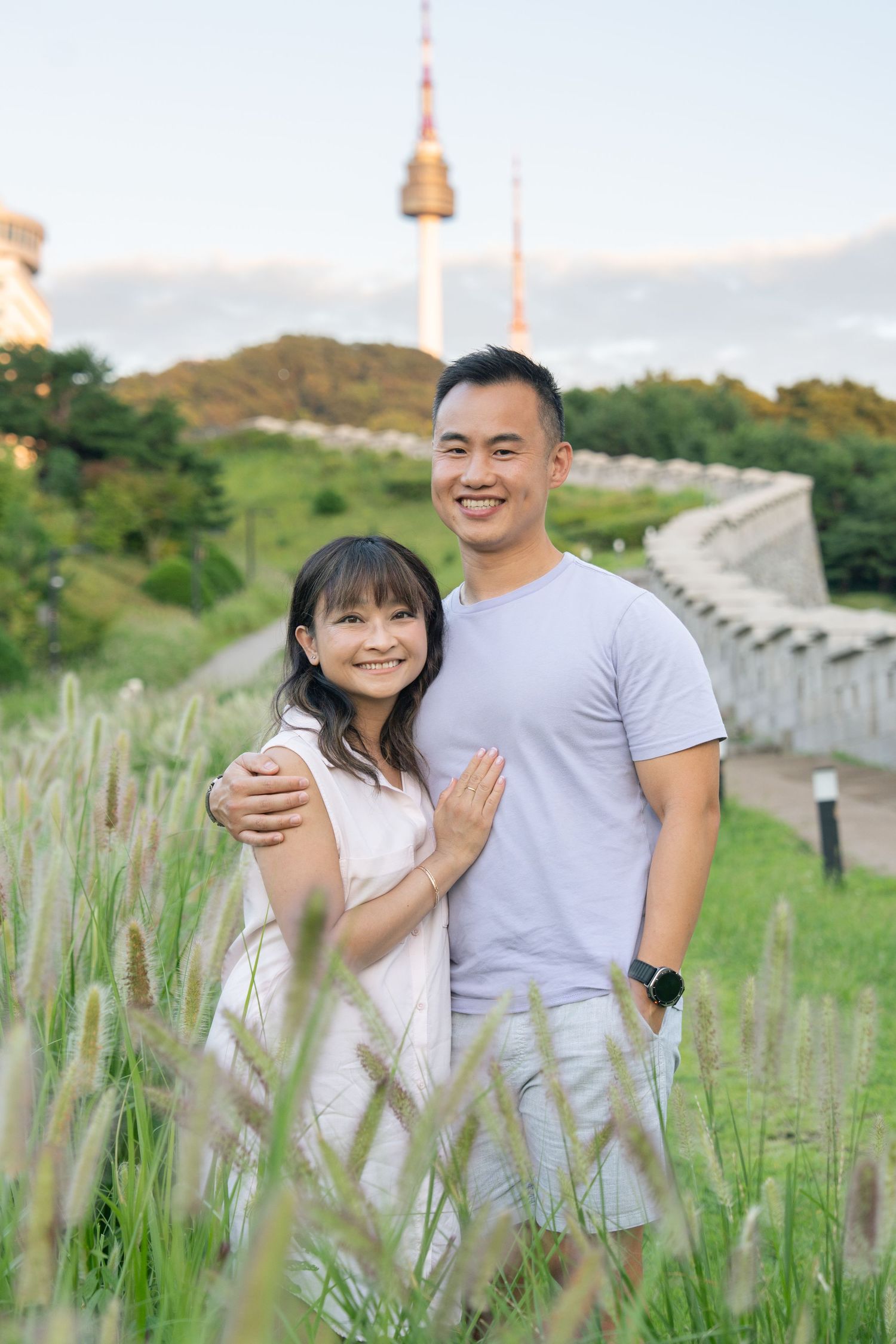 A couple embraces in a field of tall grass with a stone wall in the background during golden hour.