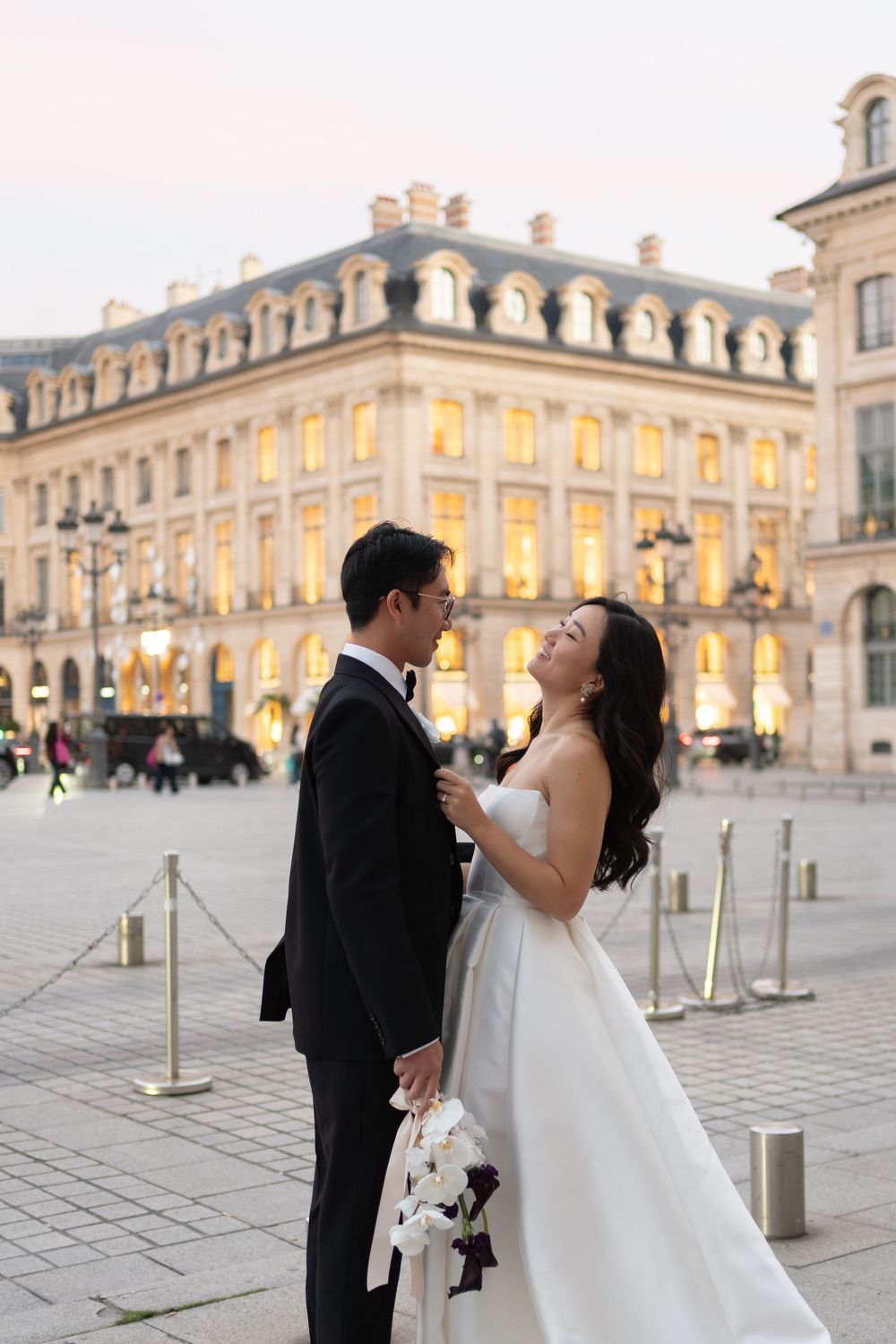 A couple in wedding attire poses romantically at dusk in Paris's Place Vendôme, with illuminated buildings behind them.
