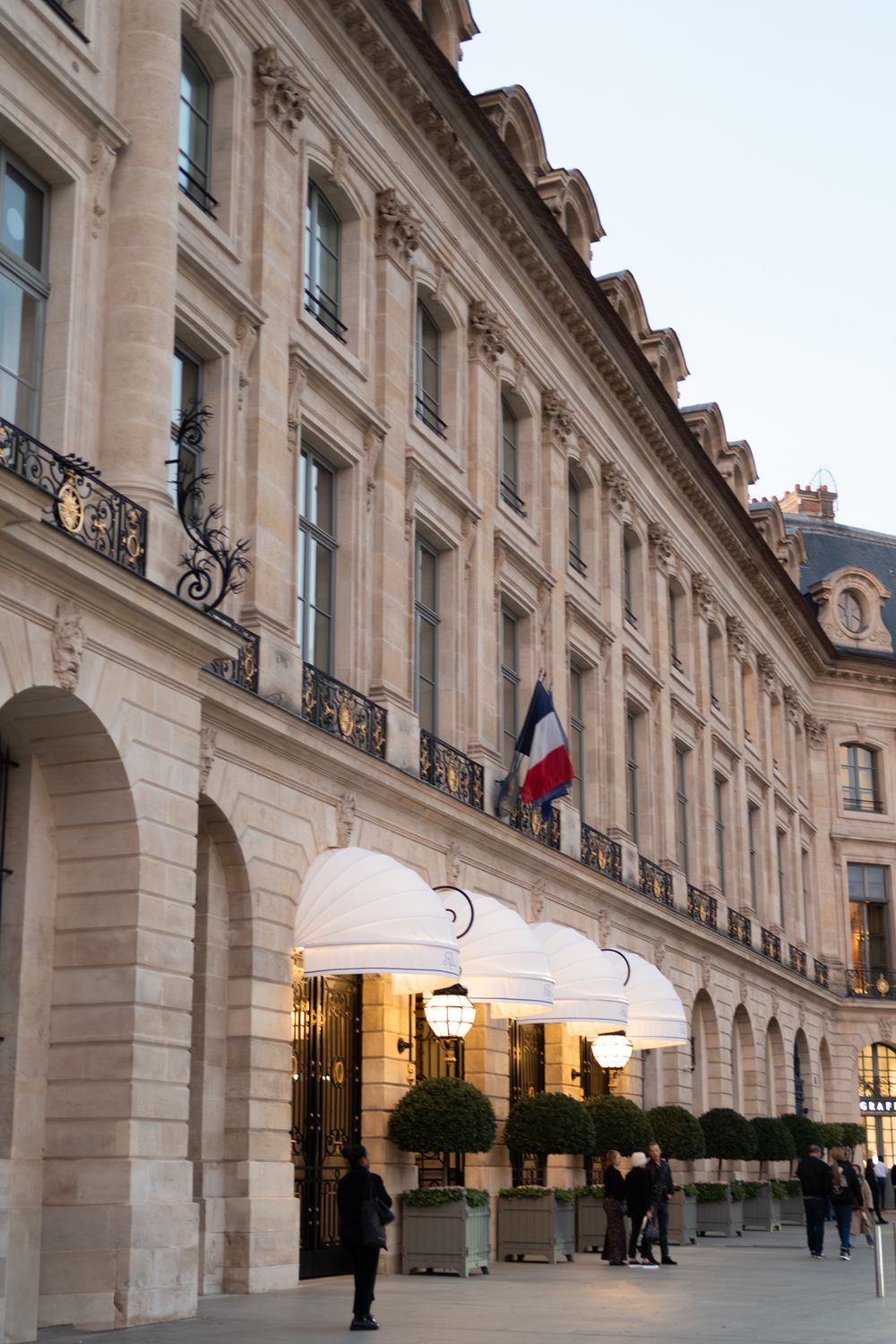Elegant stone colonnade of the Ritz Hotel in Paris with French flags and ornate architectural details illuminated at dusk.