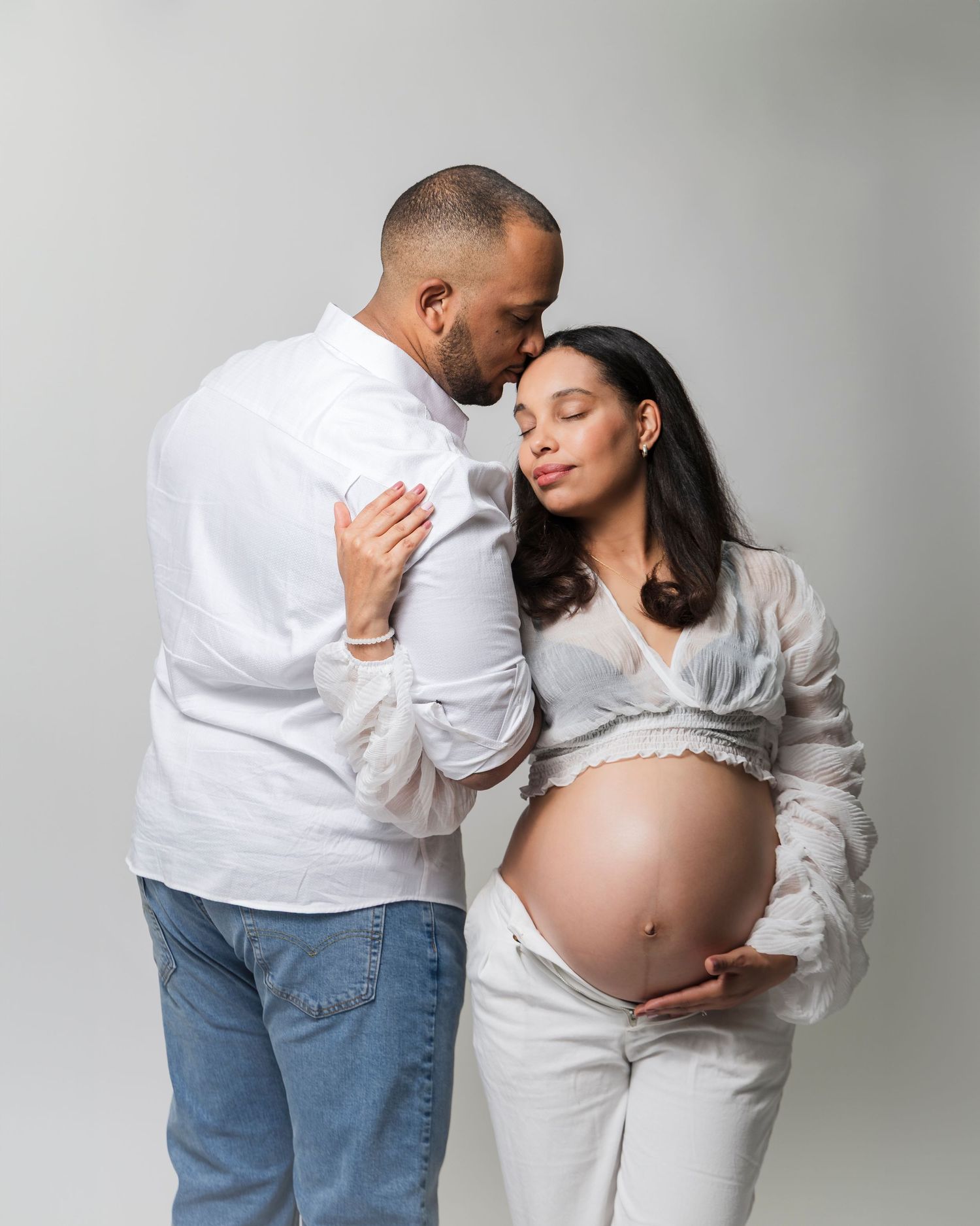 Intimate maternity photoshoot featuring couple in white and denim embracing against neutral backdrop.