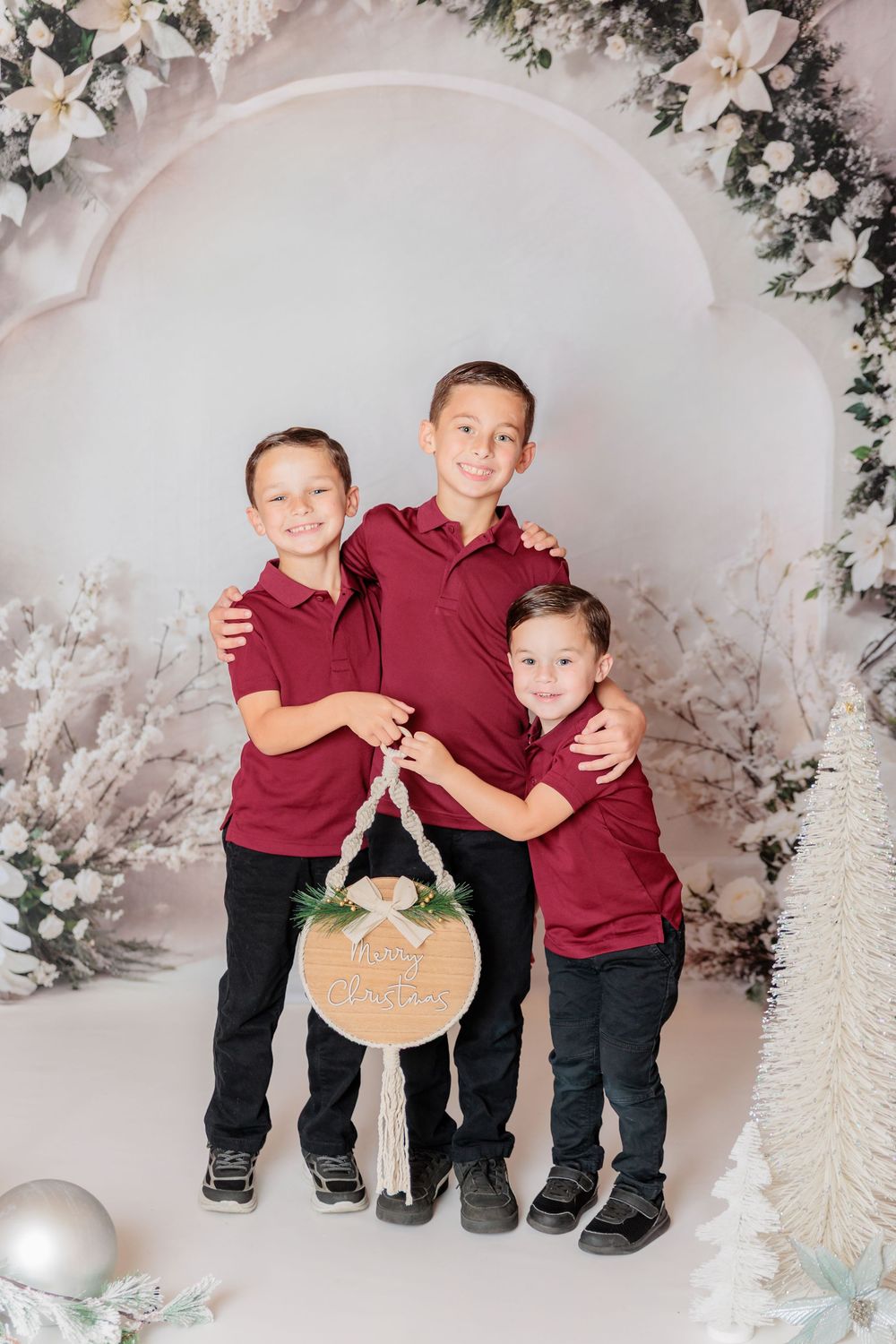 Three young children in burgundy polo shirts and dark pants pose together in a winter wonderland photo setting. Clayton NJ Photo Studio