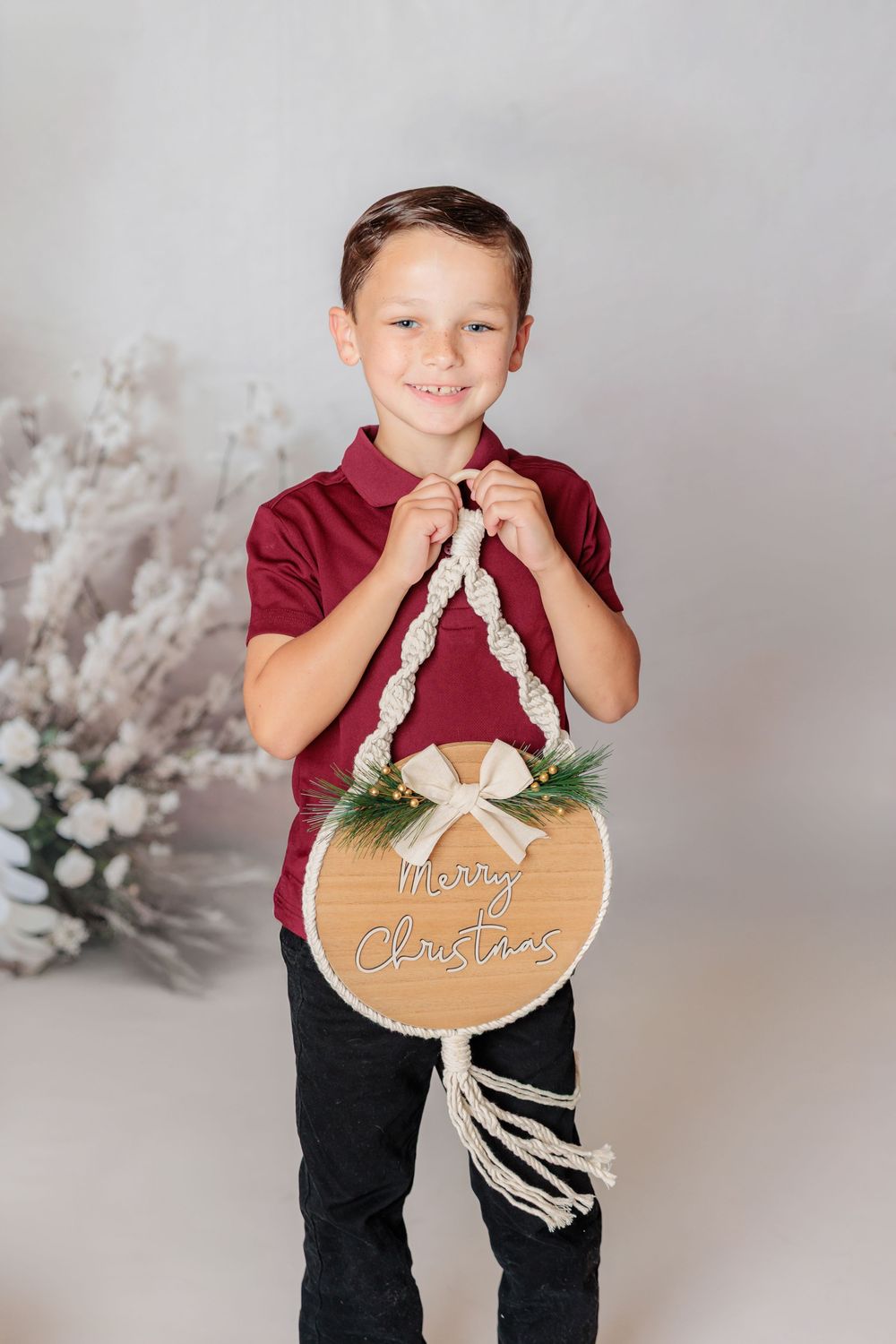 Child in burgundy shirt holds wooden Christmas ornament with white bow against grey backdrop with winter decor. Clayton NJ Photo Studio