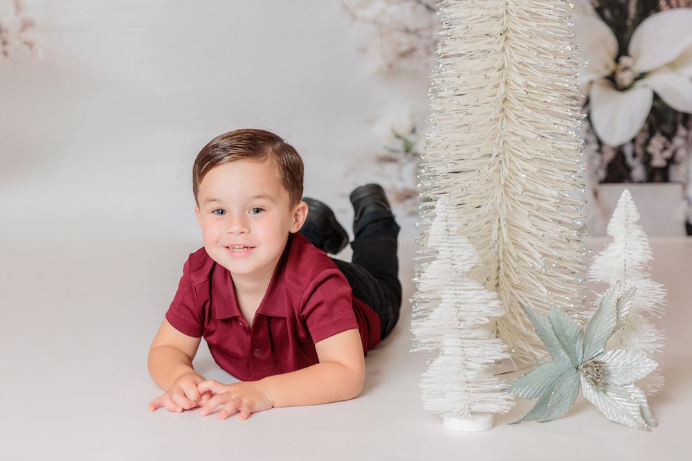 child in a burgundy shirt lies on their stomach on a white backdrop with decorative winter trees in the background. Clayton NJ Photo Studio