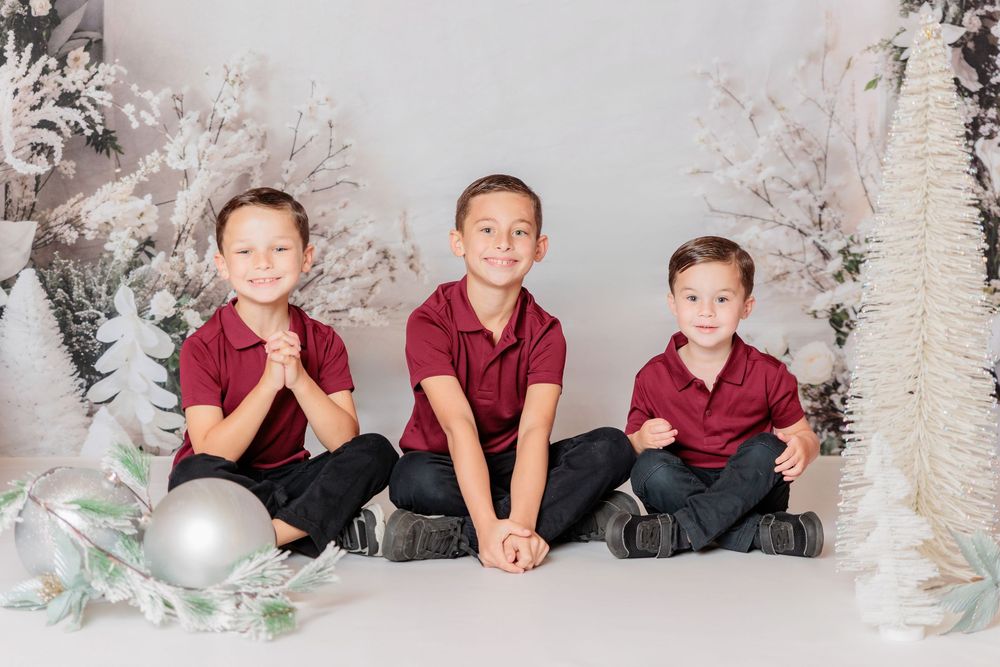 Three children in matching burgundy shirts pose together in a winter wonderland setting with trees and decorations. Clayton NJ Photo Studio