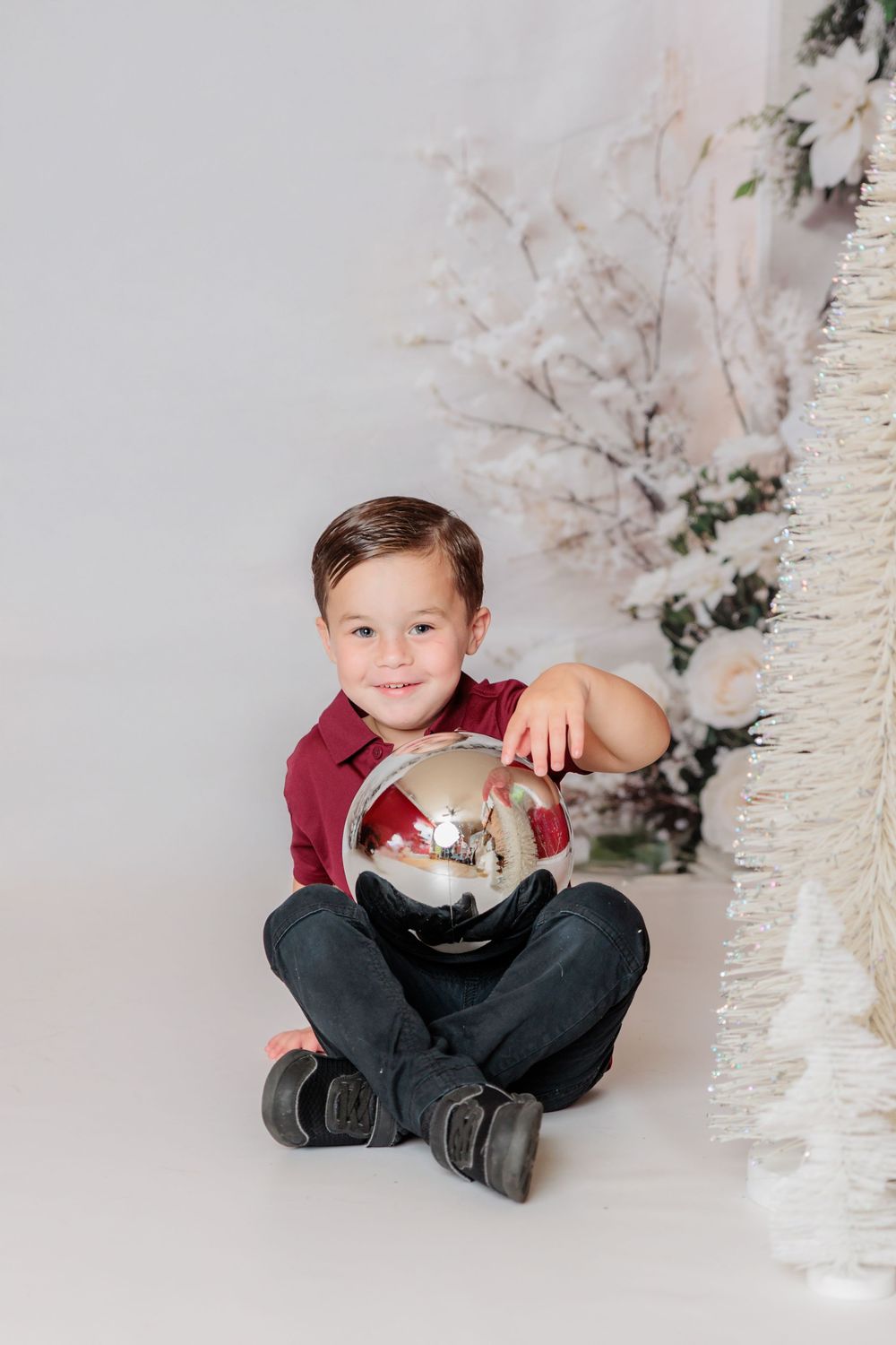 Child in burgundy shirt sits cross-legged on floor surrounded by white Christmas trees and decorations. Clayton NJ Photo Studio