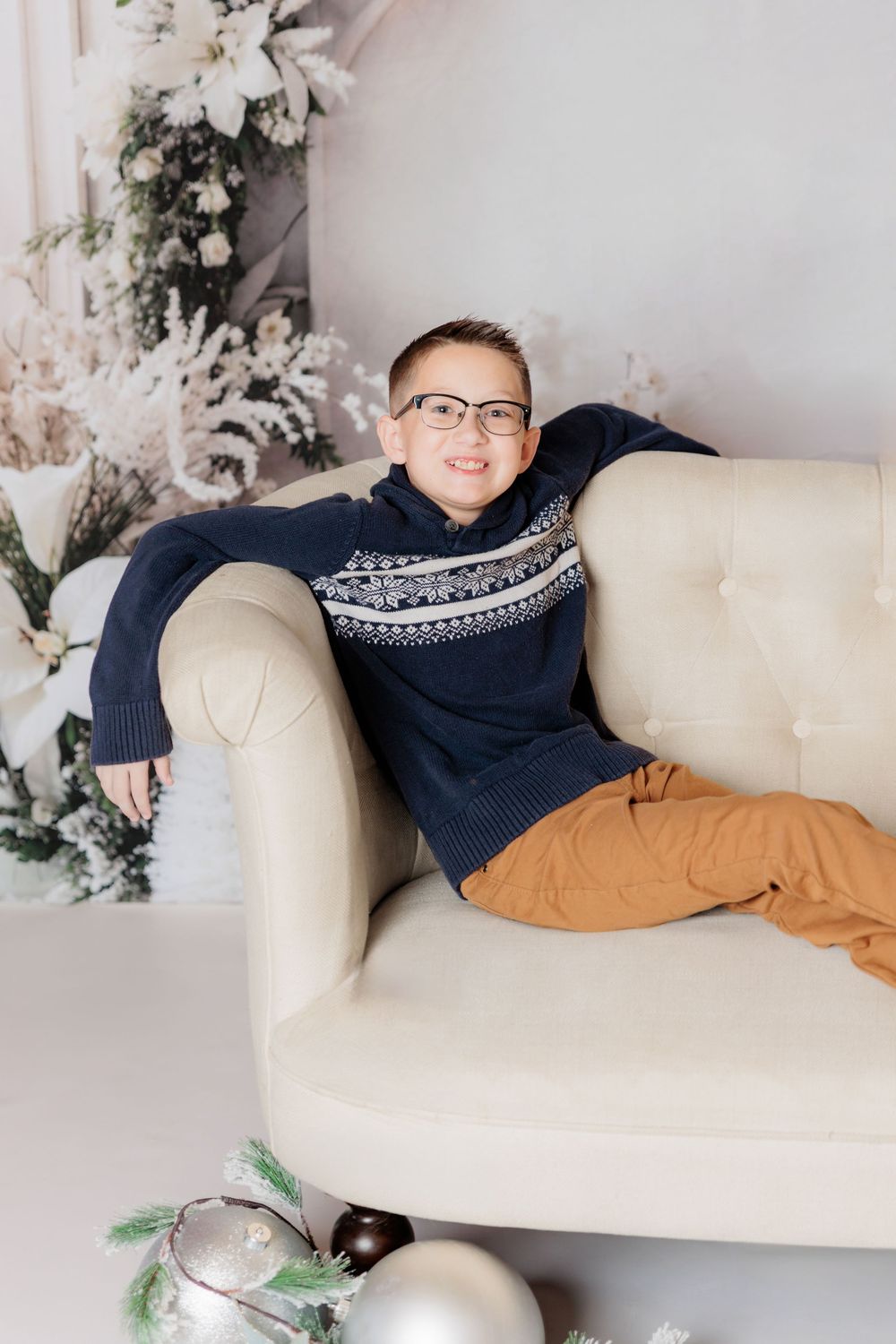 Person lounging on cream colored couch wearing navy sweater and tan pants in holiday photo setting. Clayton NJ Photo Studio