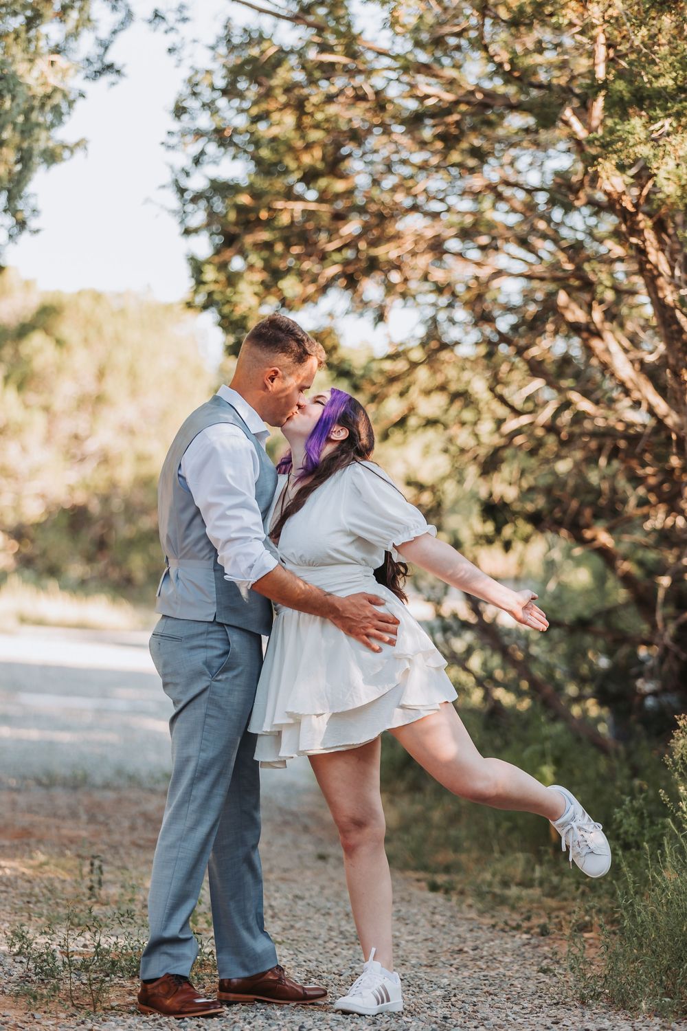 A couple shares a lighthearted dancing moment on a rustic trail with sunlit trees in the background.
