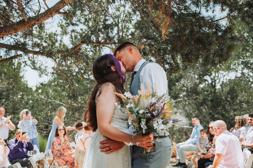 A romantic outdoor wedding ceremony under trees with guests seated on wooden chairs and a floral bouquet.
