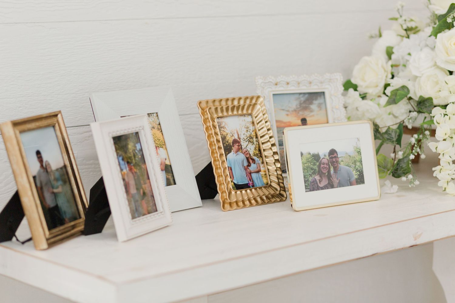 Decorative photo frames with gold and white borders displayed on a white mantle beside white roses.