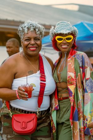 Two friends in summer outfits smile and pose together at an outdoor event wearing colorful clothing and accessories.