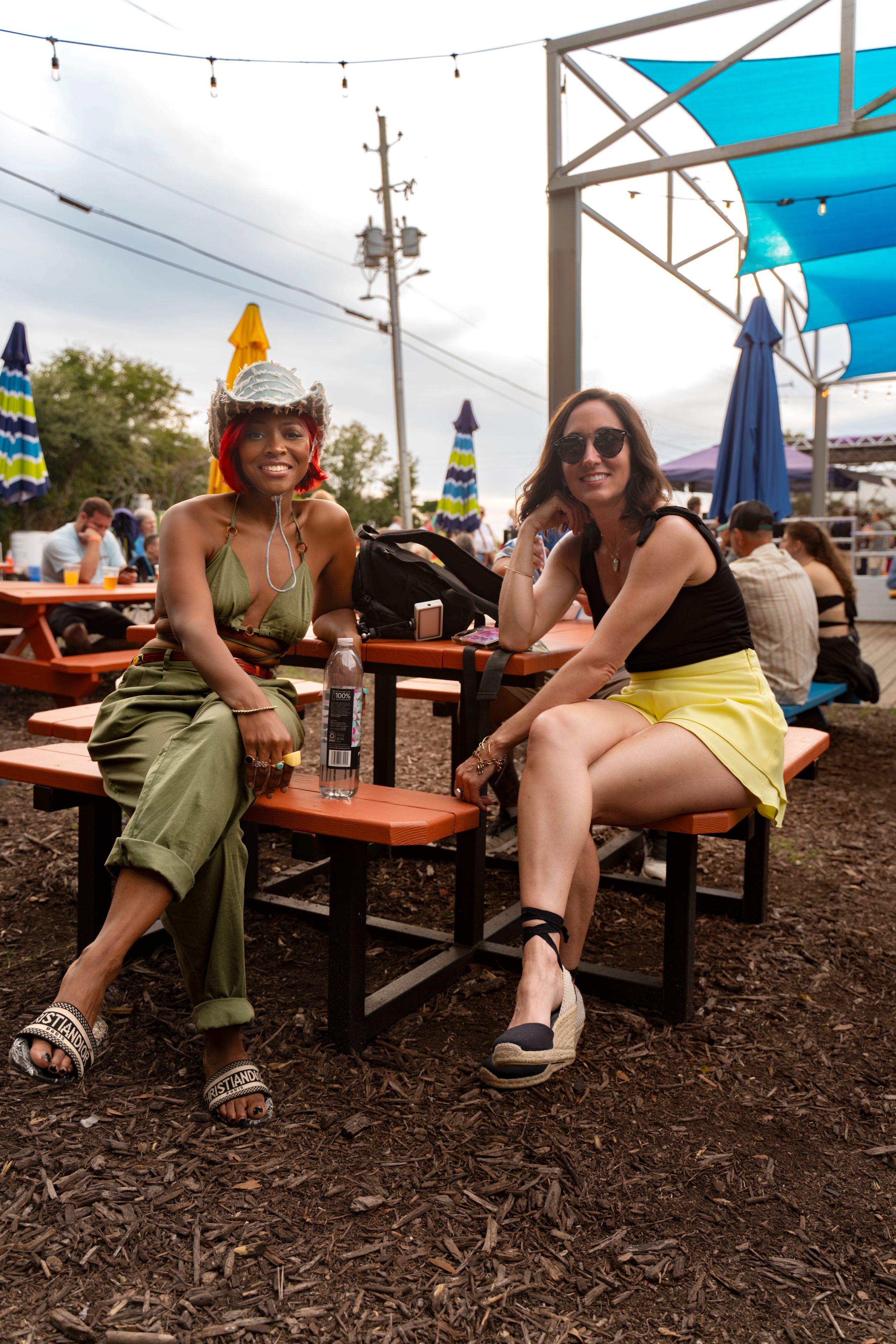 People relaxing at outdoor picnic tables on mulch ground under blue canopy on sunny day.