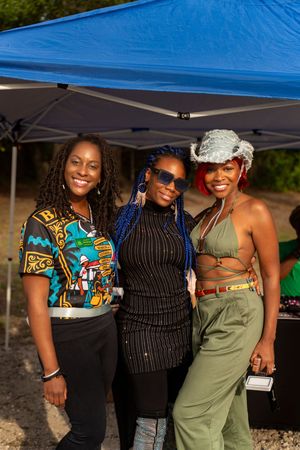 Three friends pose together under a blue canopy tent at an outdoor festival wearing colorful African prints and casual attire.