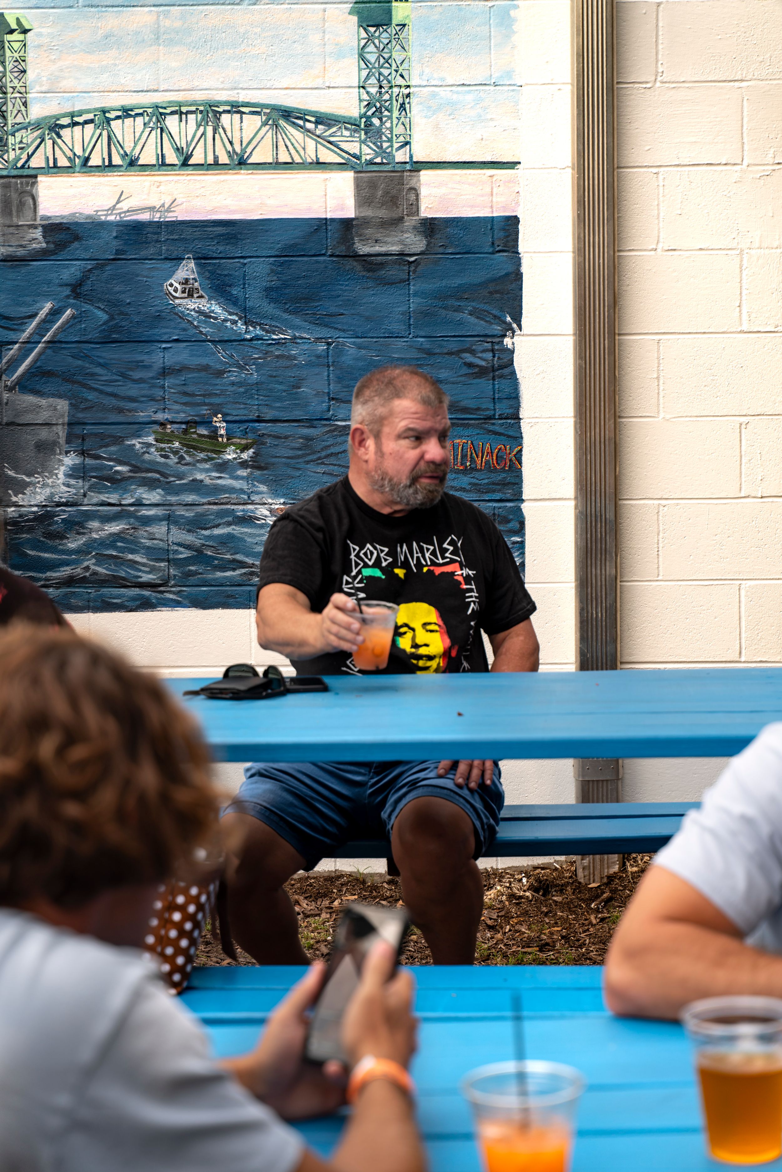 People sitting at blue picnic tables with drinks against a wall painted with an ocean mural.