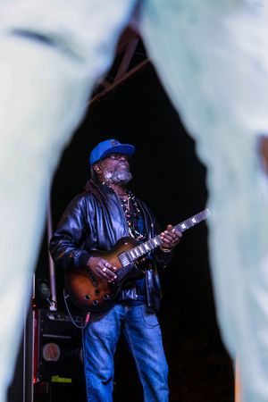 Blues musician performs on stage wearing denim and a cap while playing guitar under dramatic lighting.