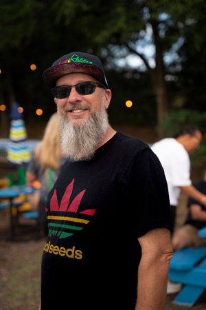 A bearded man in sunglasses and a black t-shirt smiles at an outdoor festival.
