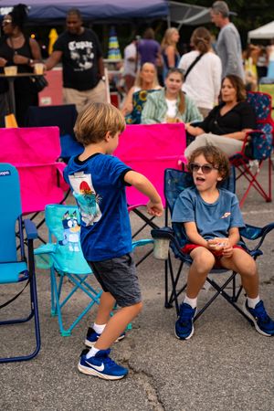 Children in blue clothing playing and moving around folding chairs at an outdoor event.