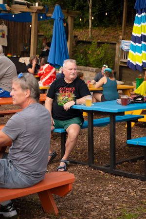 People sitting at colorful picnic tables with blue umbrellas at an outdoor venue.