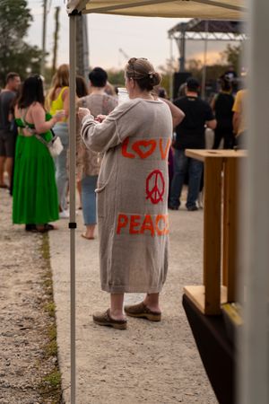 People gather outdoors under a canopy tent with someone wearing a coat displaying a peace symbol in red paint.