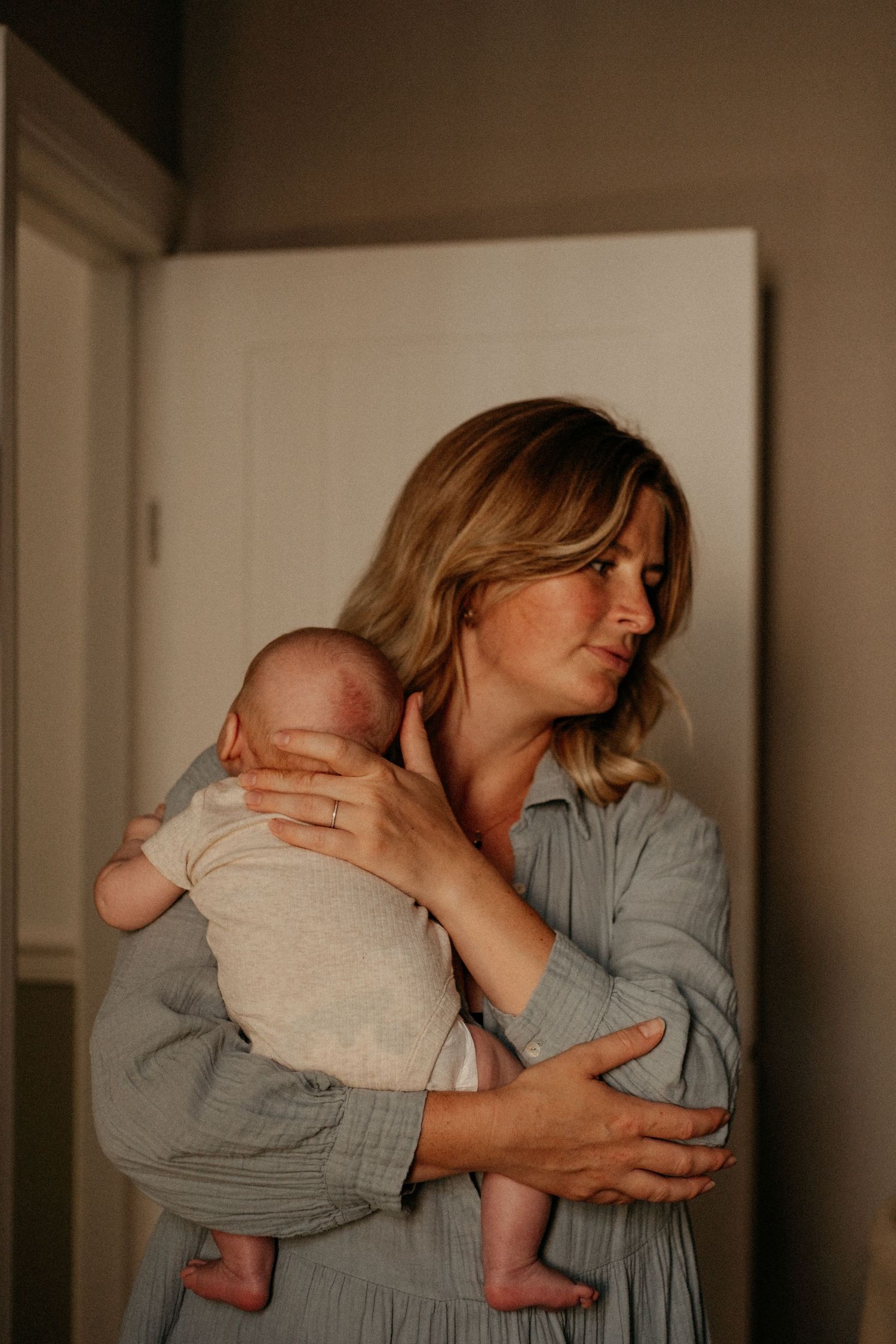 Mother holding her newborn baby in a softly lit doorway during an intimate in-home motherhood photography session.