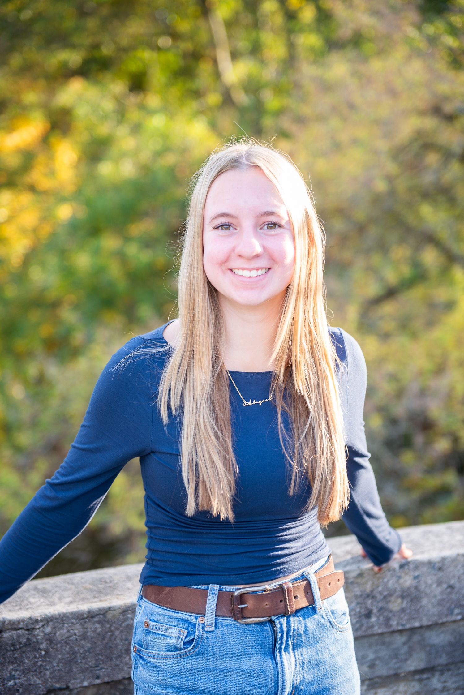 Person in navy blue top and jeans poses casually against stone wall outdoors with autumn foliage in background.