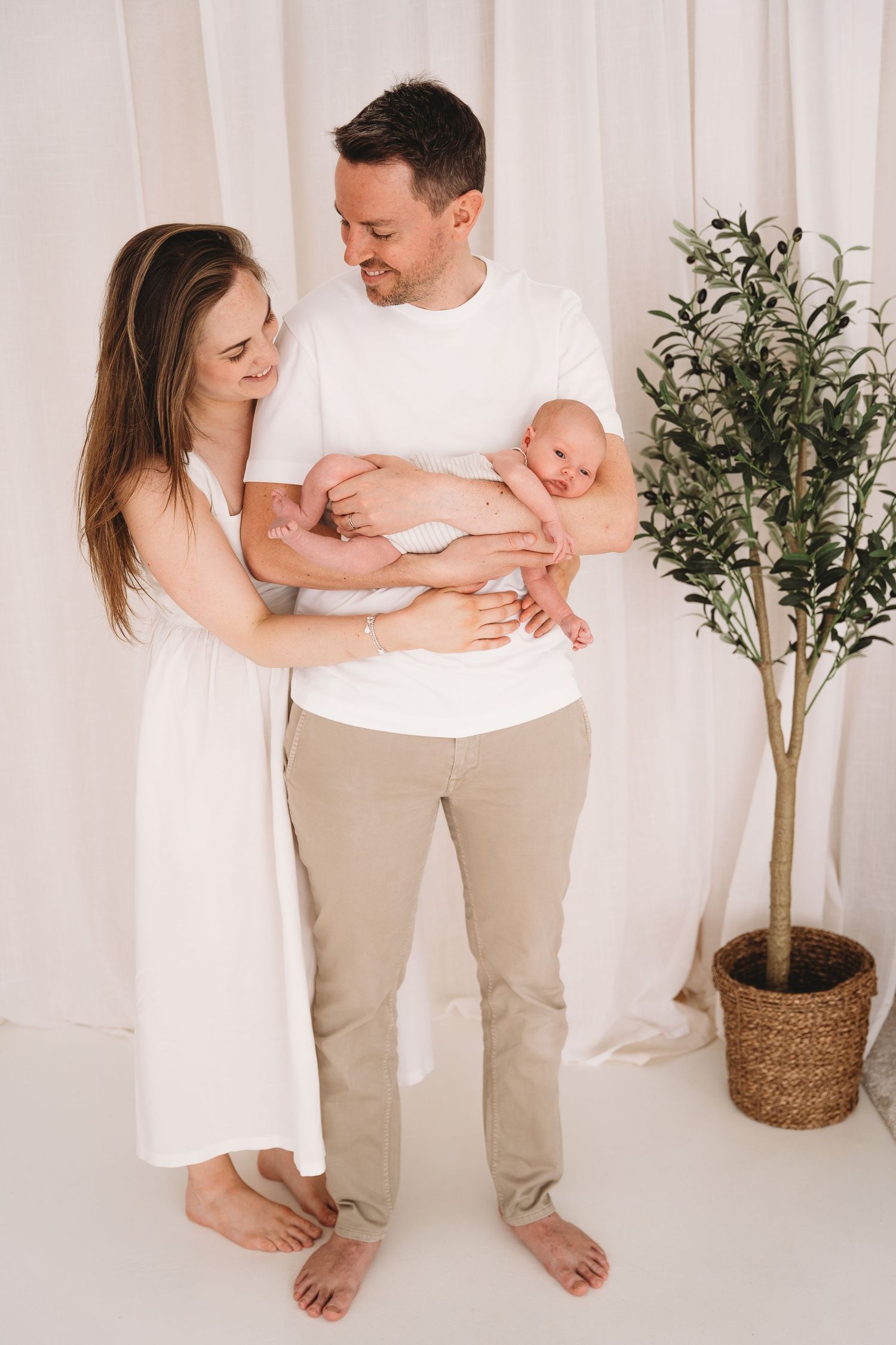 Mama en papa in lichte kledij houden hun baby vast en lachen naar elkaar.