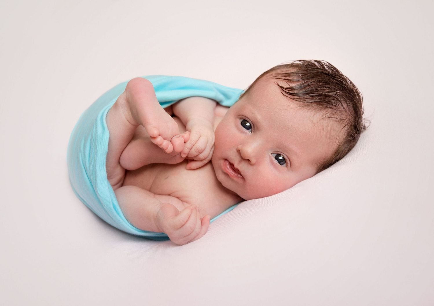 A newborn baby in light blue clothing lying on a soft white backdrop in various peaceful sleeping poses.