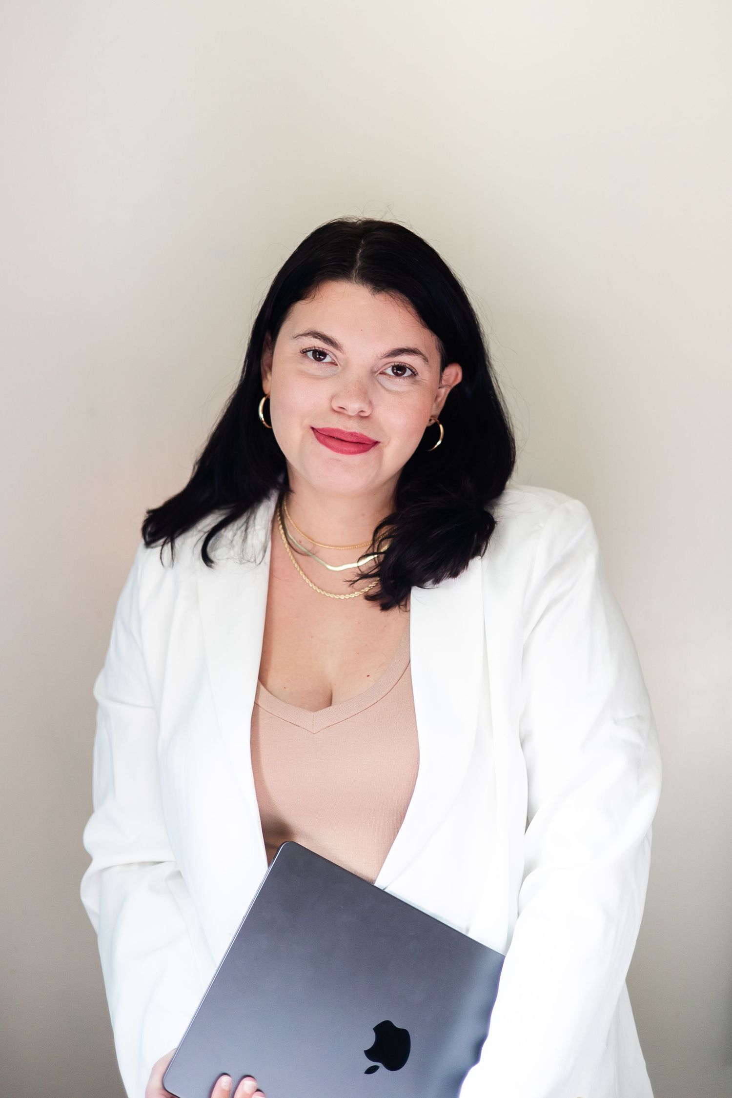 Professional photographer wearing a white blazer and tan bodysuit with gold jewelry poses with a laptop against a neutral background.