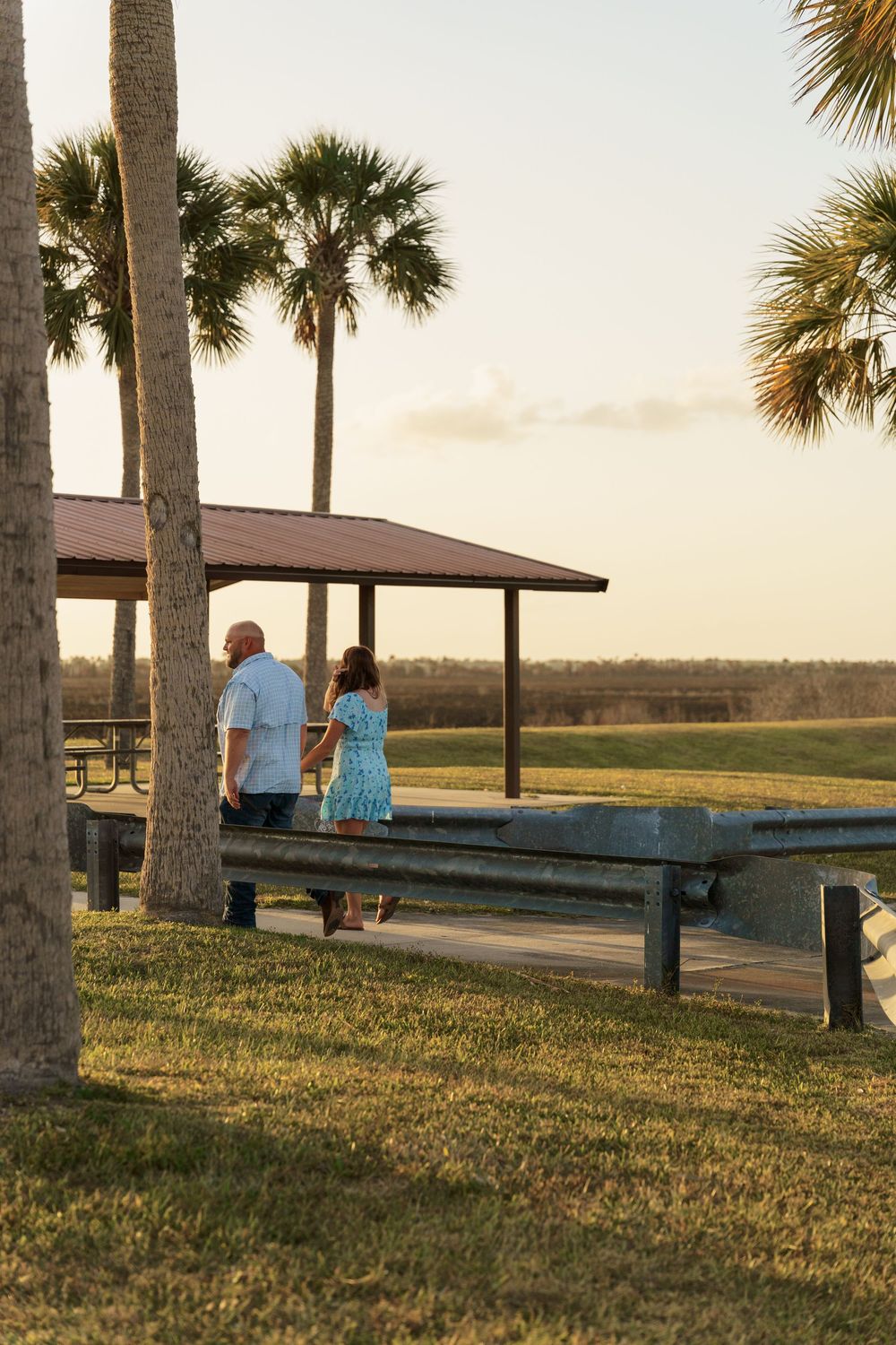 A couple sits on a bench at sunset overlooking a grassy coastal area with palm trees framing the peaceful scene.