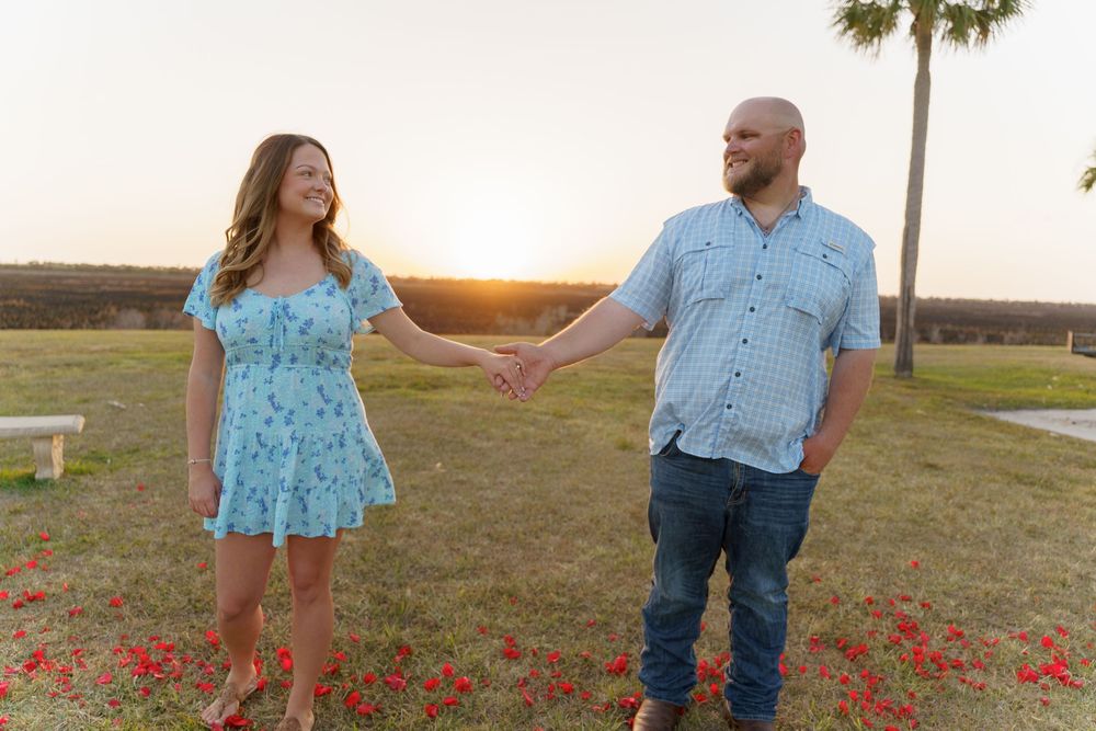 A couple in blue outfits holds hands while walking through a field of red wildflowers at sunset.