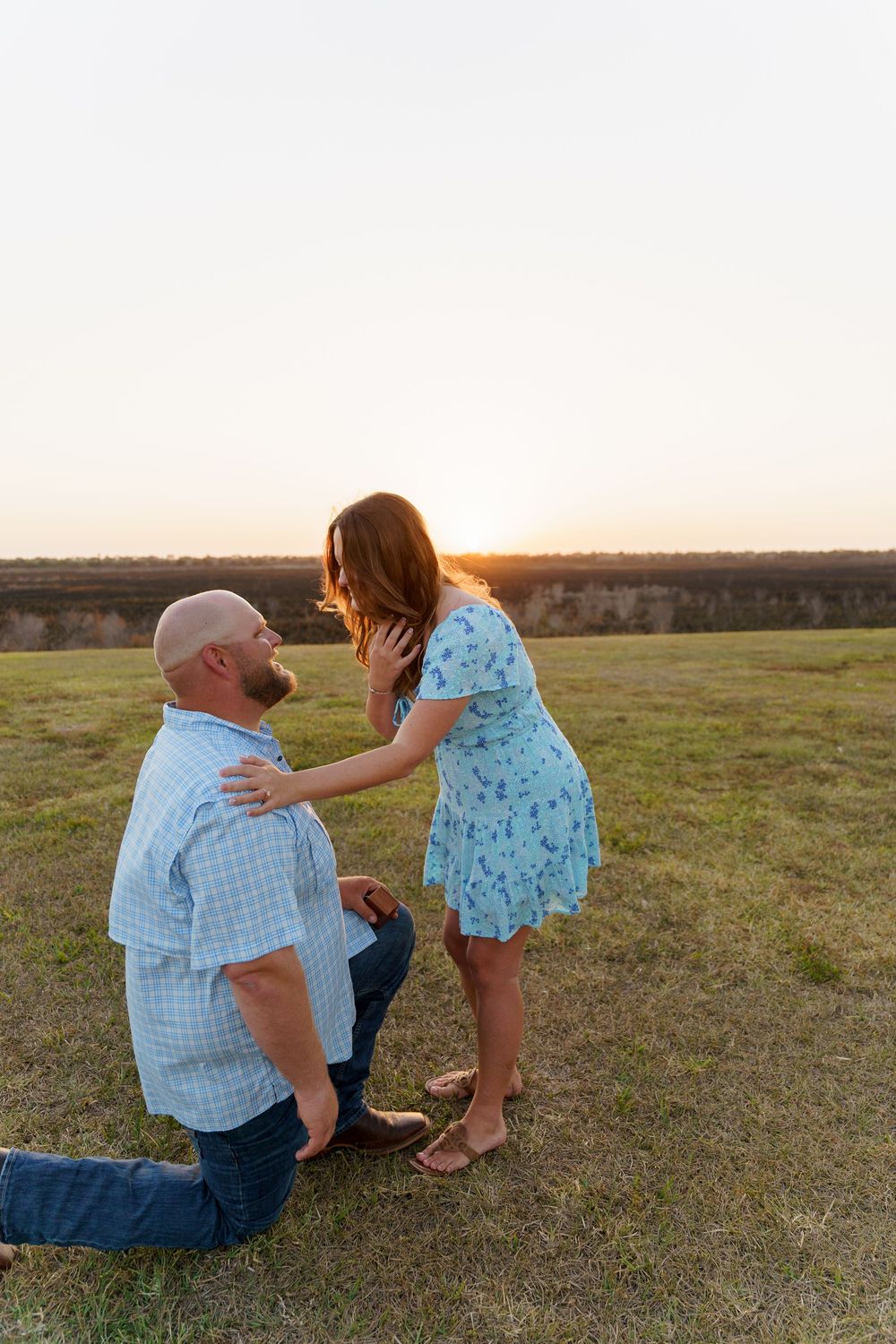 A couple shares a romantic moment during sunset in a field as one kneels before the other in a blue floral dress.
