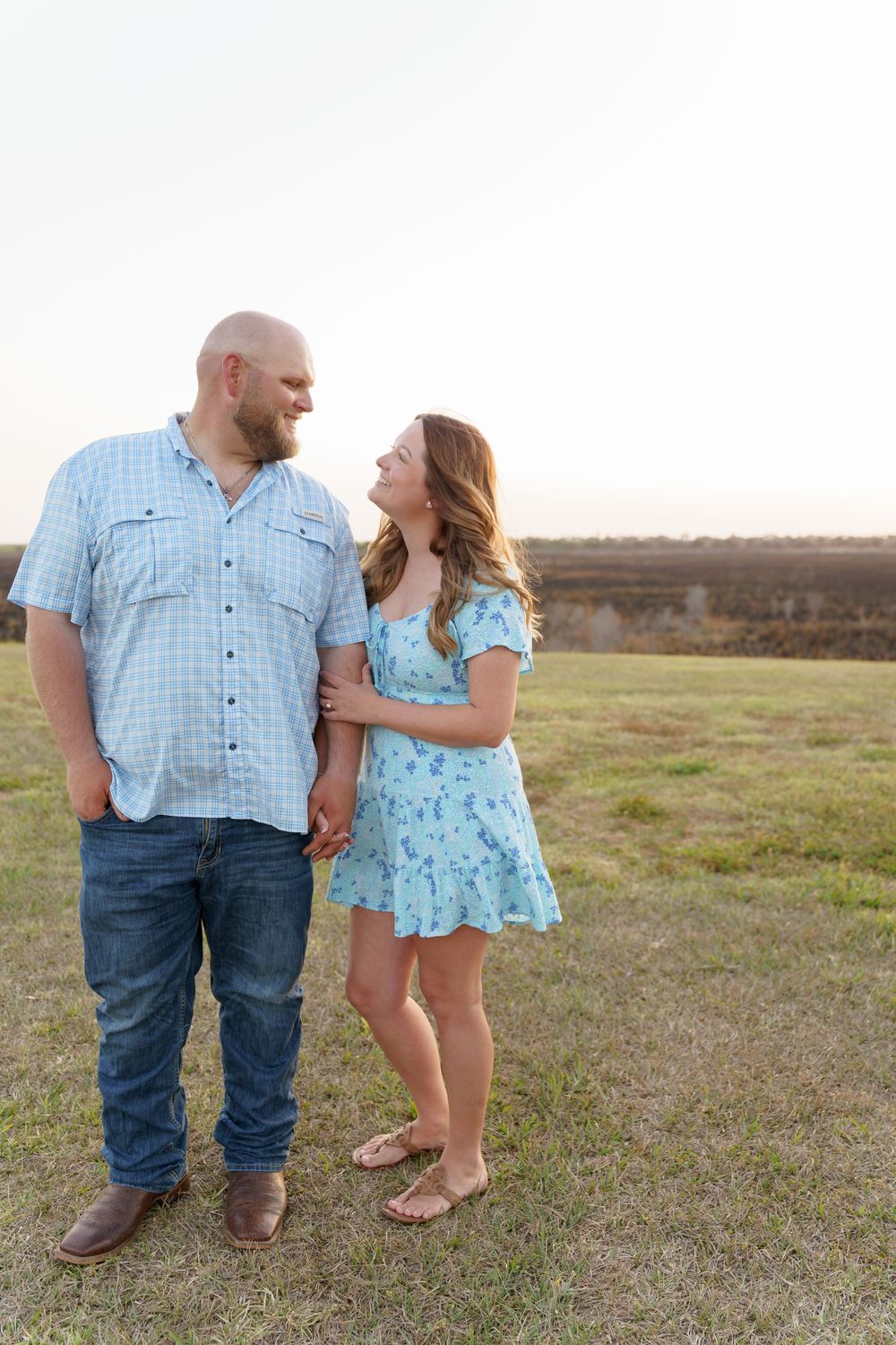 A young girl in a blue floral dress stands with someone in jeans and a plaid shirt in a grassy field at sunset.