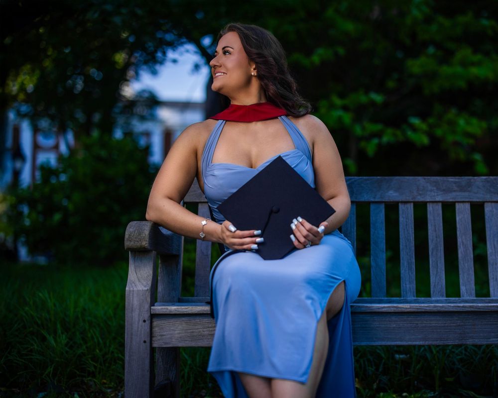 A person in blue and black clothing poses on a wooden bench in a lush garden setting during twilight.