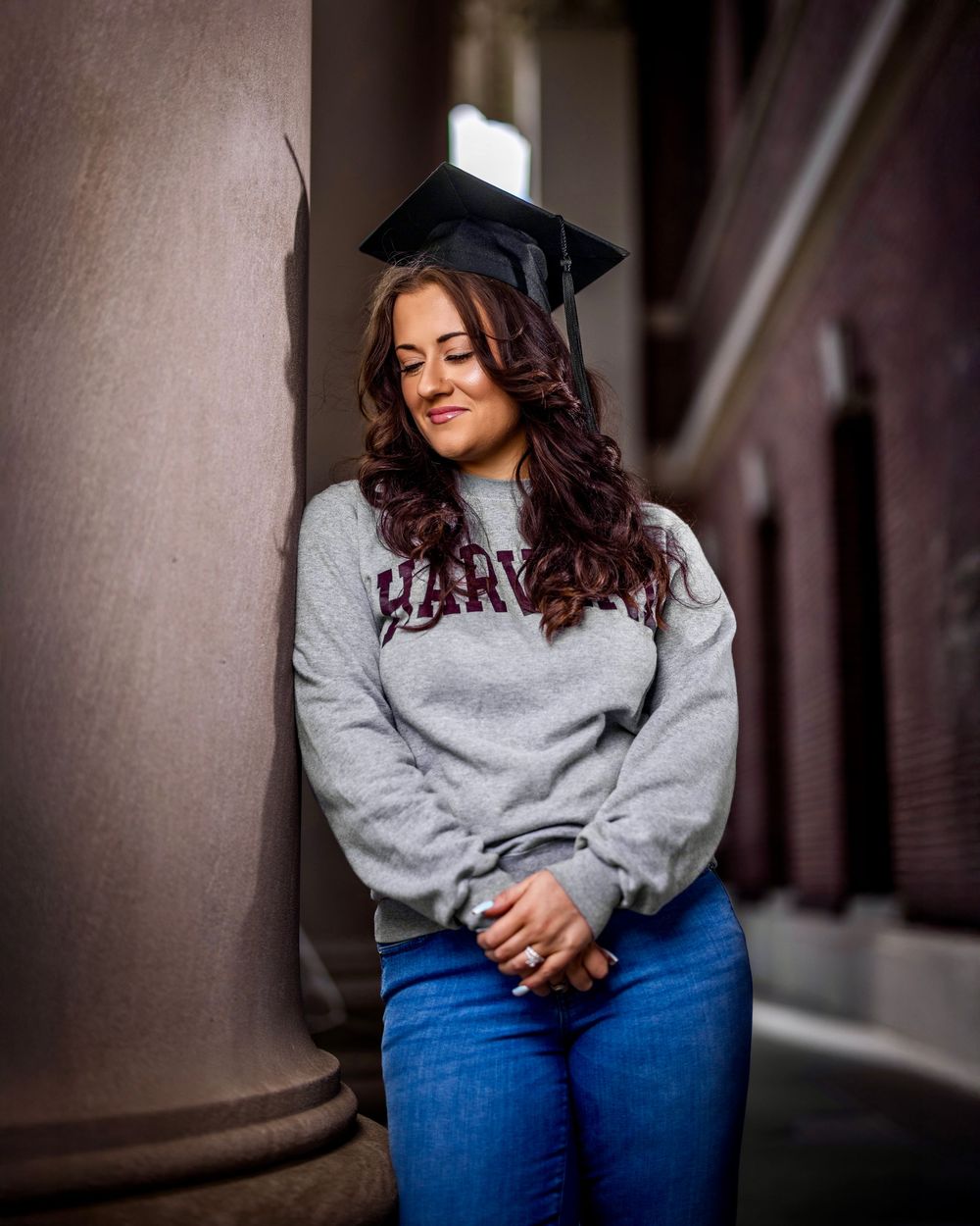 Graduate wearing a Harvard sweatshirt and graduation cap leans against a stone pillar in a contemplative pose.