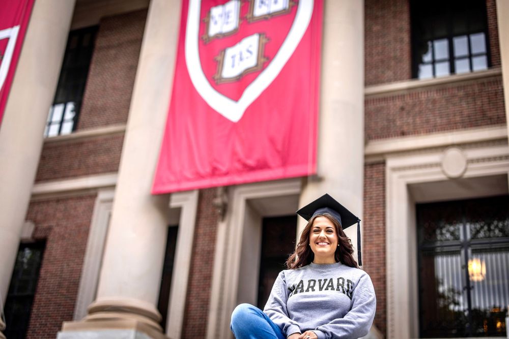 Graduates in caps and gowns celebrate on campus with pink banners and brick buildings in the background.