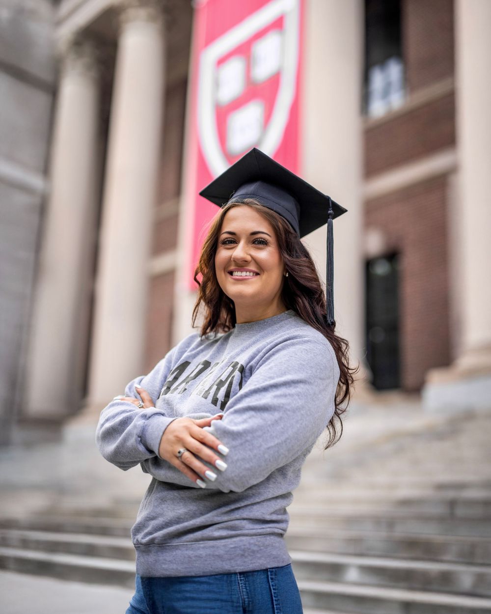 Graduate in a grey sweatshirt and graduation cap poses joyfully on campus steps.