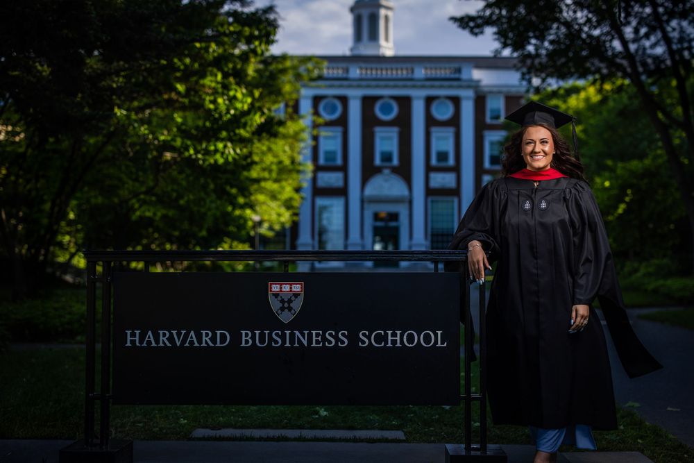 Graduate stands in academic regalia in front of Harvard Business School sign and building on campus.