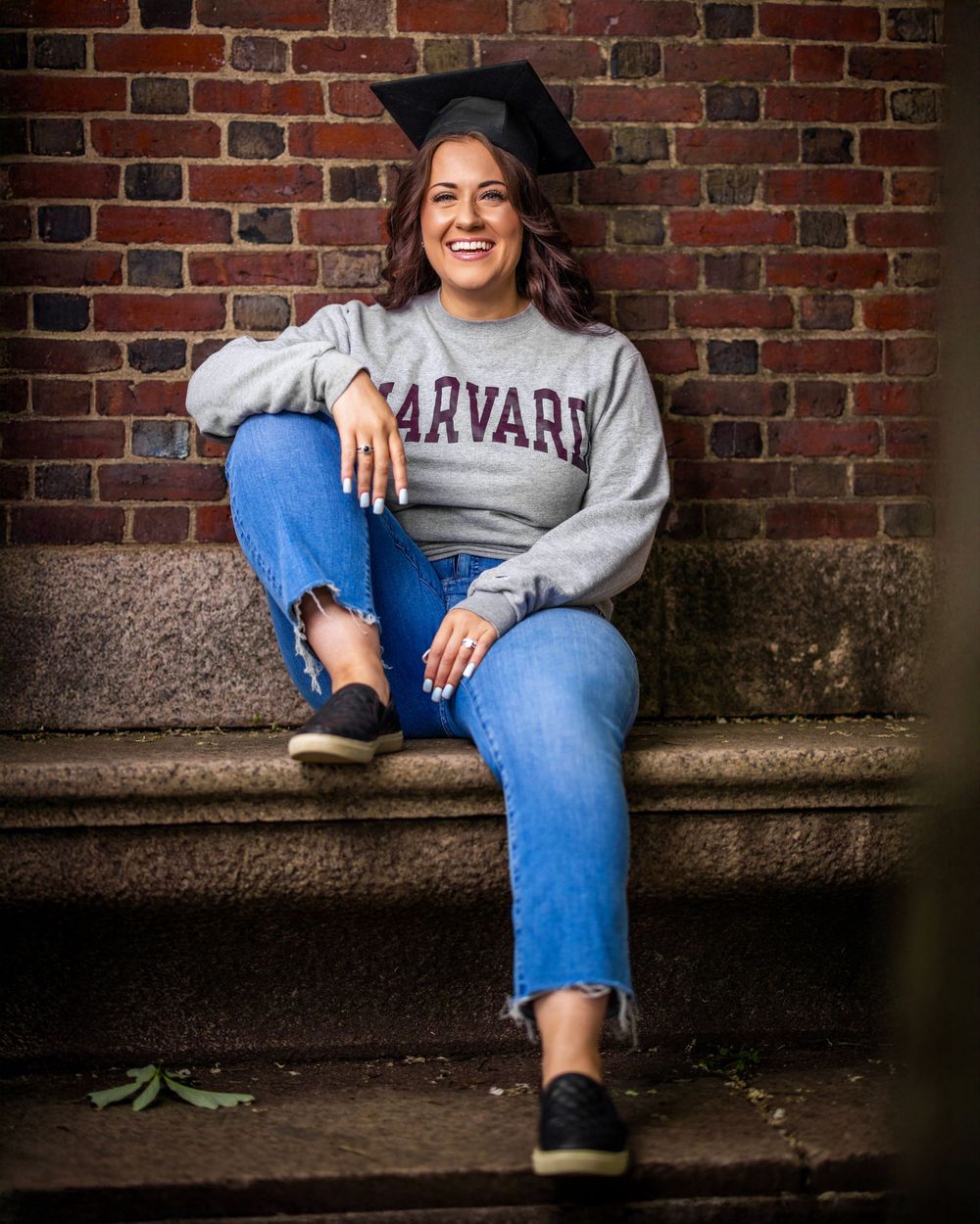 A student in a Harvard sweatshirt and jeans sits casually on a stone bench against a brick wall backdrop.