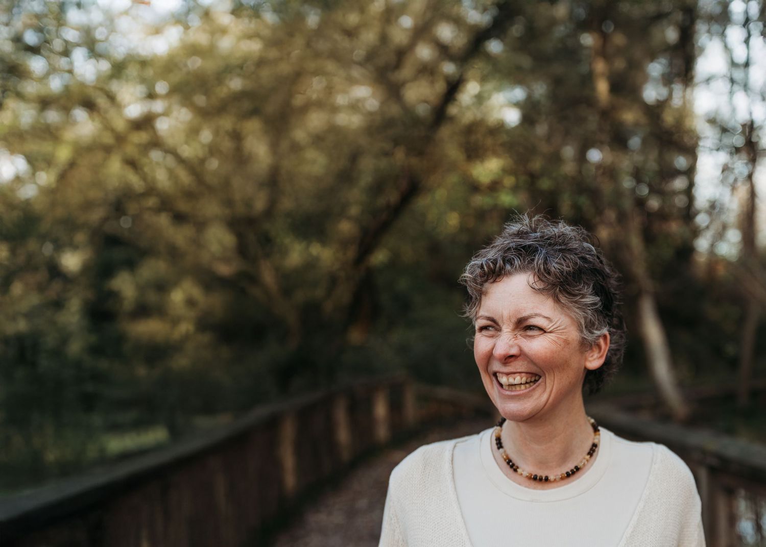 A person in a white dress smiles joyfully while standing in a wooded outdoor setting during golden hour.